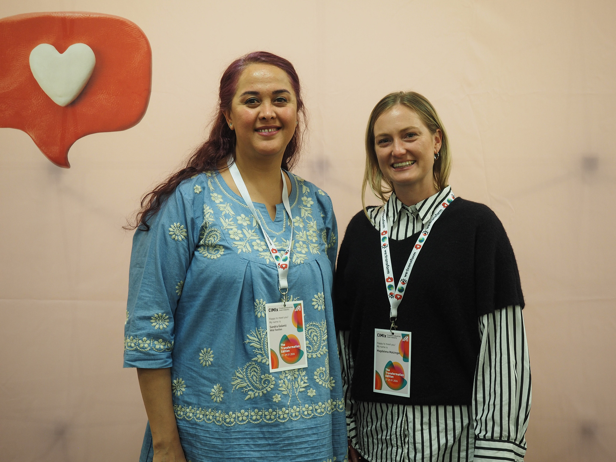Zwei Frauen mit CIMIx 2026-Konferenzabzeichen stehen nebeneinander und lächeln in die Kamera. Die Frau auf der linken Seite trägt ein hellblaues besticktes Kleid, die Frau auf der rechten Seite einen schwarzen Pullover über einem gestreiften Hemd. Hinter ihnen ist eine Herz-Sprechblase zu sehen.