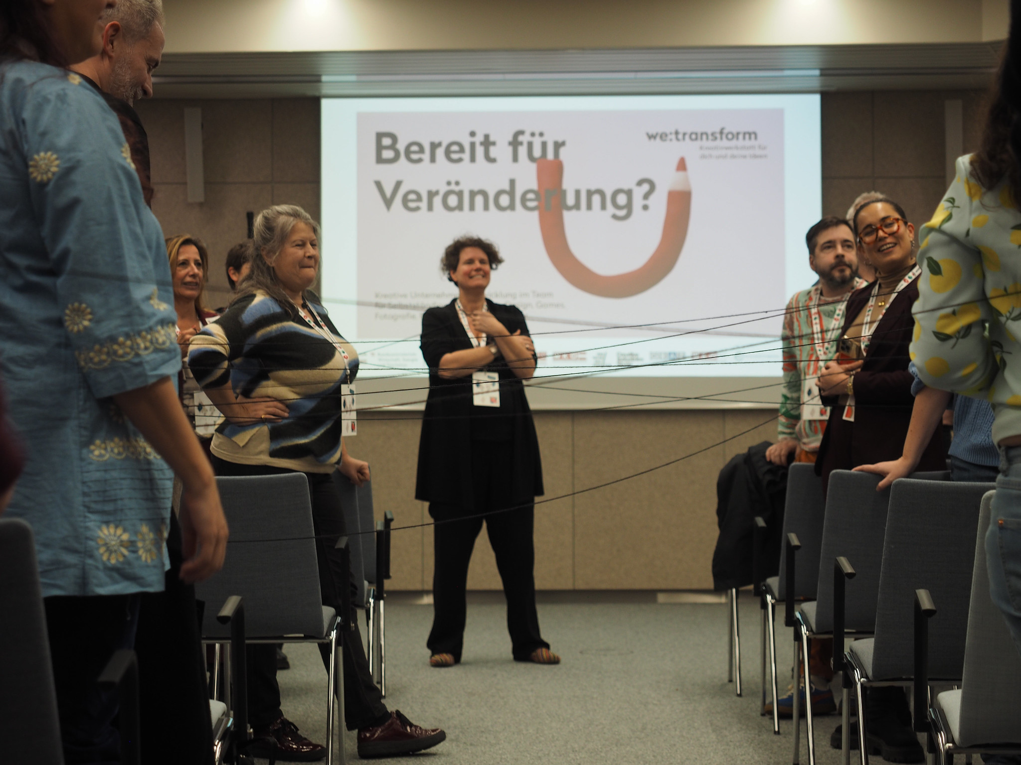 Eine Gruppe von Menschen steht sich in zwei Reihen gegenüber und hält Schnüre zwischen sich. Auf einem Bildschirm hinter ihnen erscheint der deutsche Text "Bereit für Veränderung?", das we:transform-Logo und ein Hinweis auf CIMIx 2026.