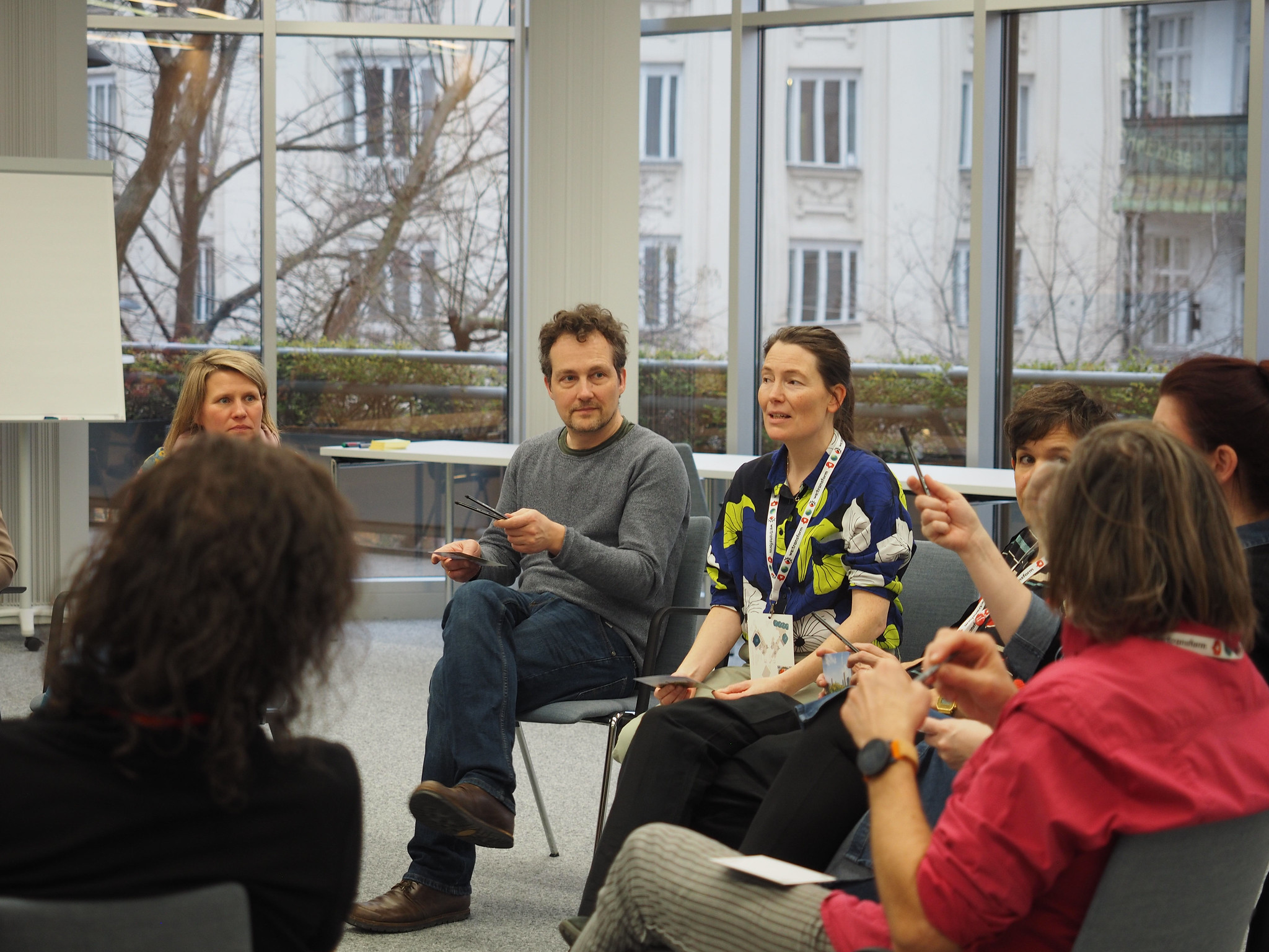 In einem hellen Raum mit großen Fenstern sitzt eine Gruppe von Menschen im Kreis und diskutiert über CIMIX 2026; einige halten Papiere in der Hand, während eine Person spricht, und andere hören aufmerksam zu.