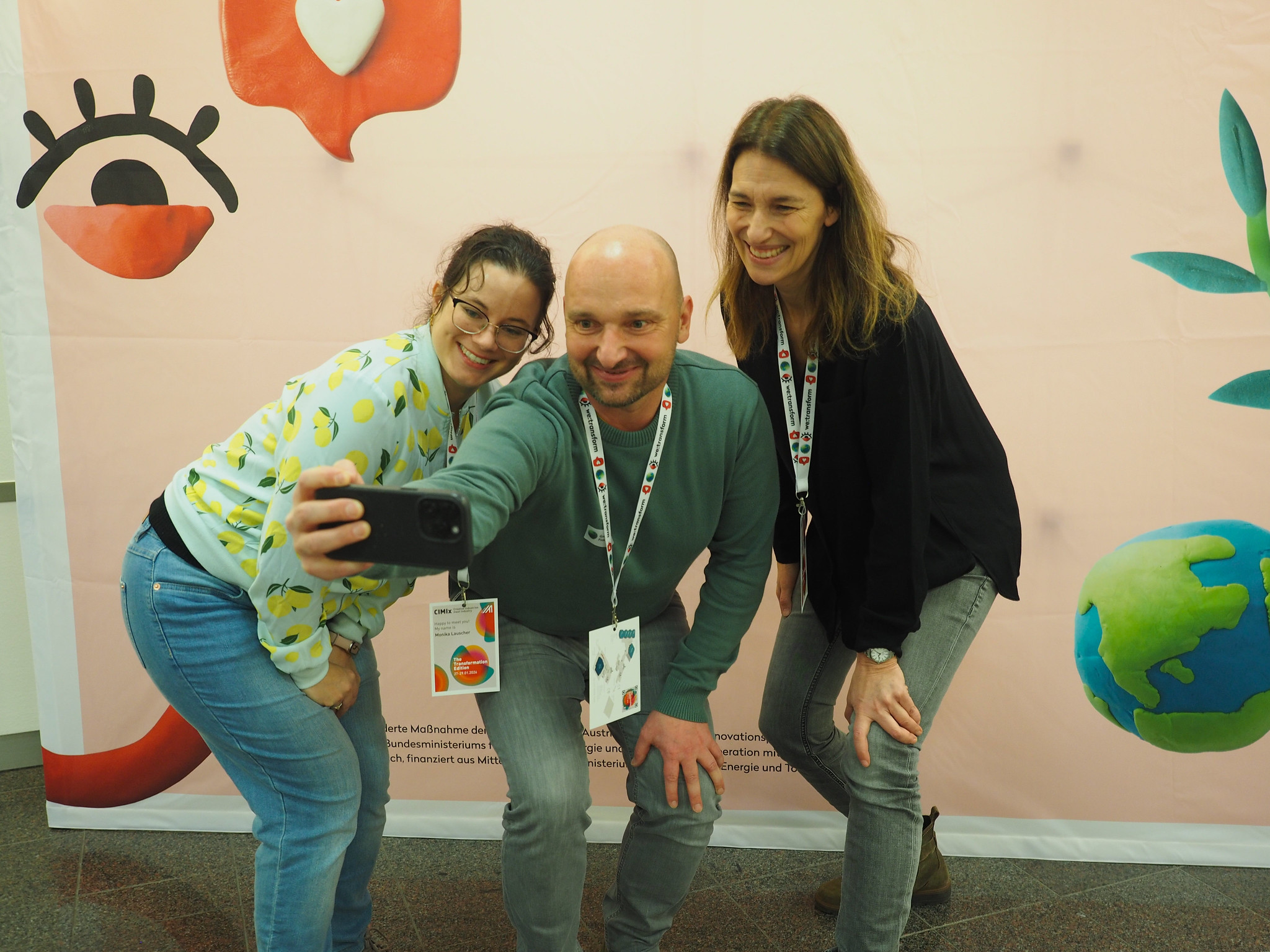 Drei Personen lächeln und posieren für ein Selfie vor einem farbenfrohen CIMIx 2026-Hintergrund mit verspielten Grafiken wie einem Auge, einer Sprechblase und einer Weltkugel. Alle tragen Konferenzabzeichen um den Hals.