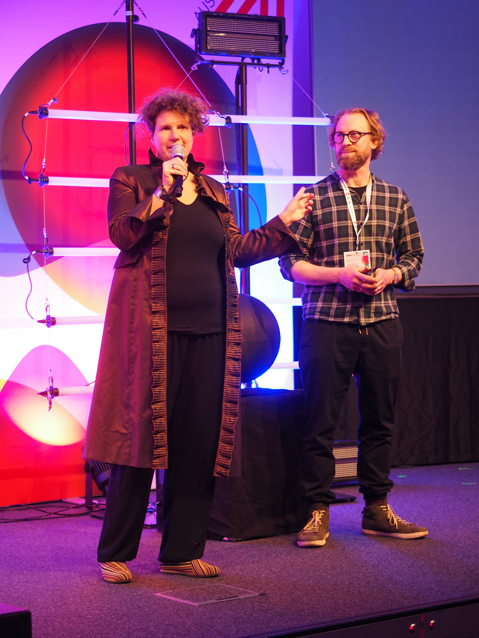 Eine Frau mit einem Mikrofon spricht auf der Bühne neben einem Mann mit einem Schlüsselband bei Connecting the Dots 2026. Im Hintergrund sind bunte Lichter, geometrische Formen und ein großer Bildschirm zu sehen. Beide sind leger gekleidet und stehen auf einem mit Teppich ausgelegten Podium.