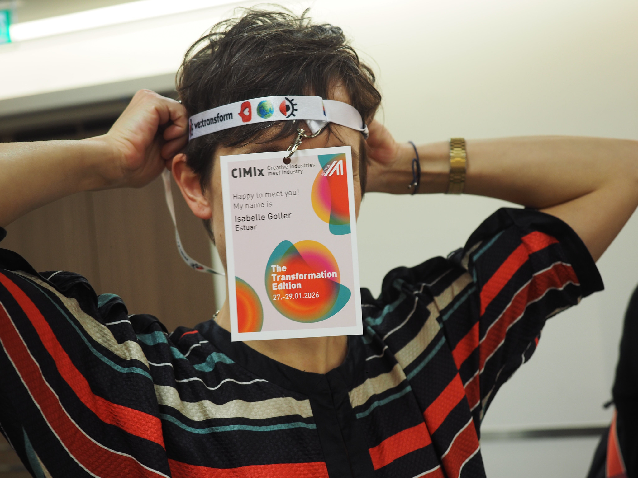 Eine Person in einem bunt gestreiften Hemd hält ein Konferenzabzeichen mit beiden Händen über ihr Gesicht. Auf dem Badge steht Isabelle Goller Estur, The Transformation Edition, 27-29.01.2020, was auf den Geist von CIMIx 2026 hinweist.