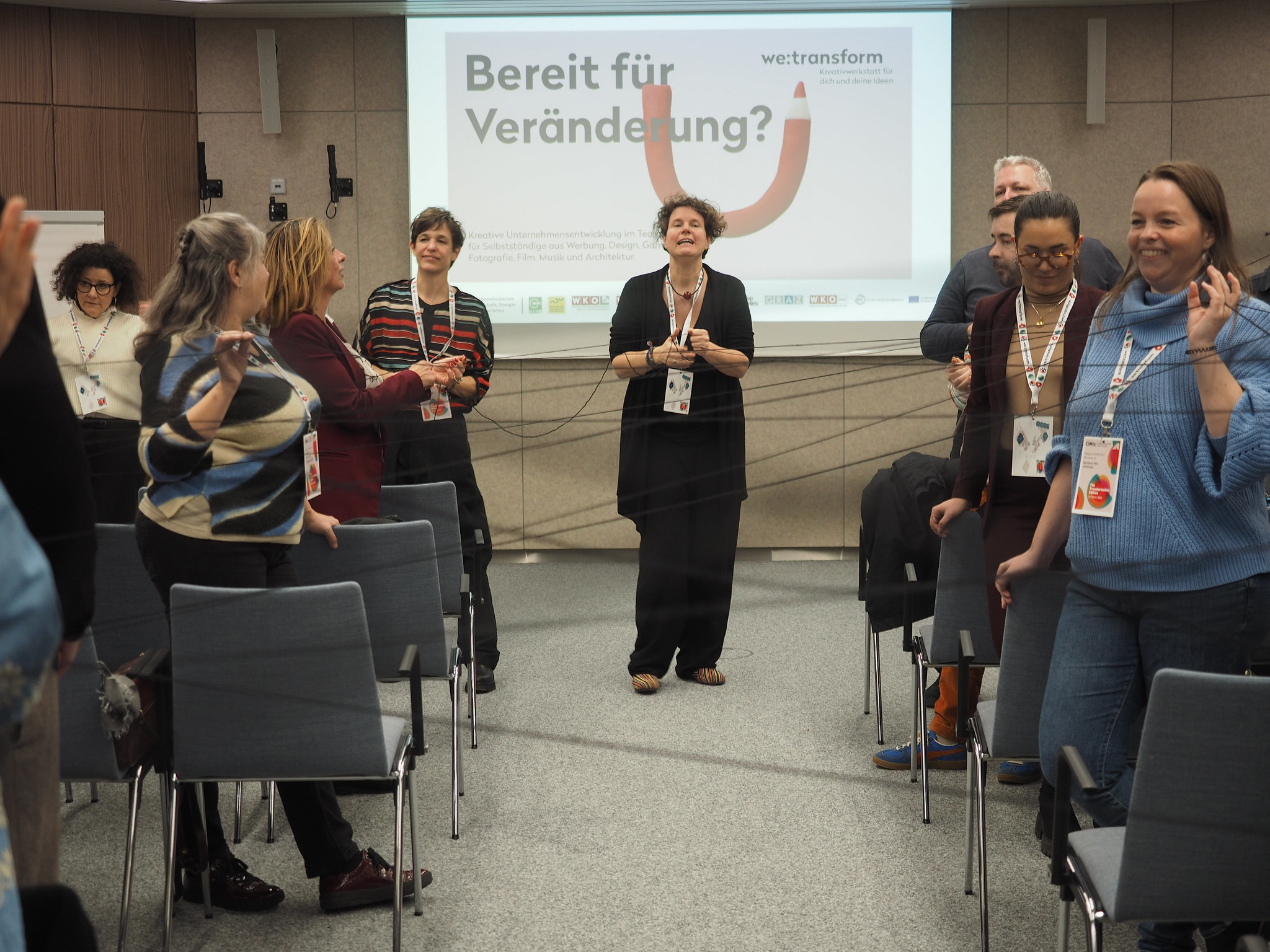 Eine Gruppe von Menschen steht in einem Konferenzraum, lächelt und unterhält sich. Eine Frau leitet die Sitzung. Auf einem Bildschirm hinter ihr steht "Bereit für Veränderung?", während sie über "Connecting the dots 2026" diskutieren.