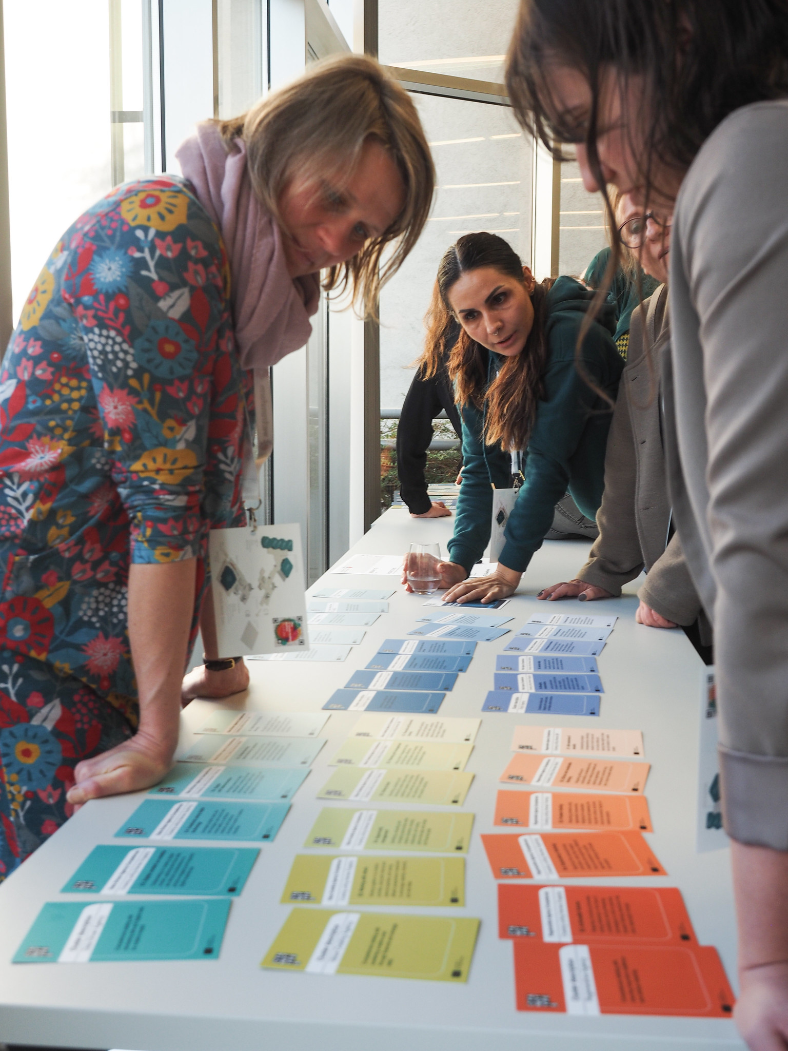 Auf der CIMIx 2026 stehen vier Personen um einen Tisch herum und betrachten bunte Karten, die in Reihen angeordnet sind. Die Karten sind blau, gelb, orange und blaugrün, jede mit einem aufgedruckten Text. Die Umgebung scheint ein heller Innenraum mit großen Fenstern zu sein.