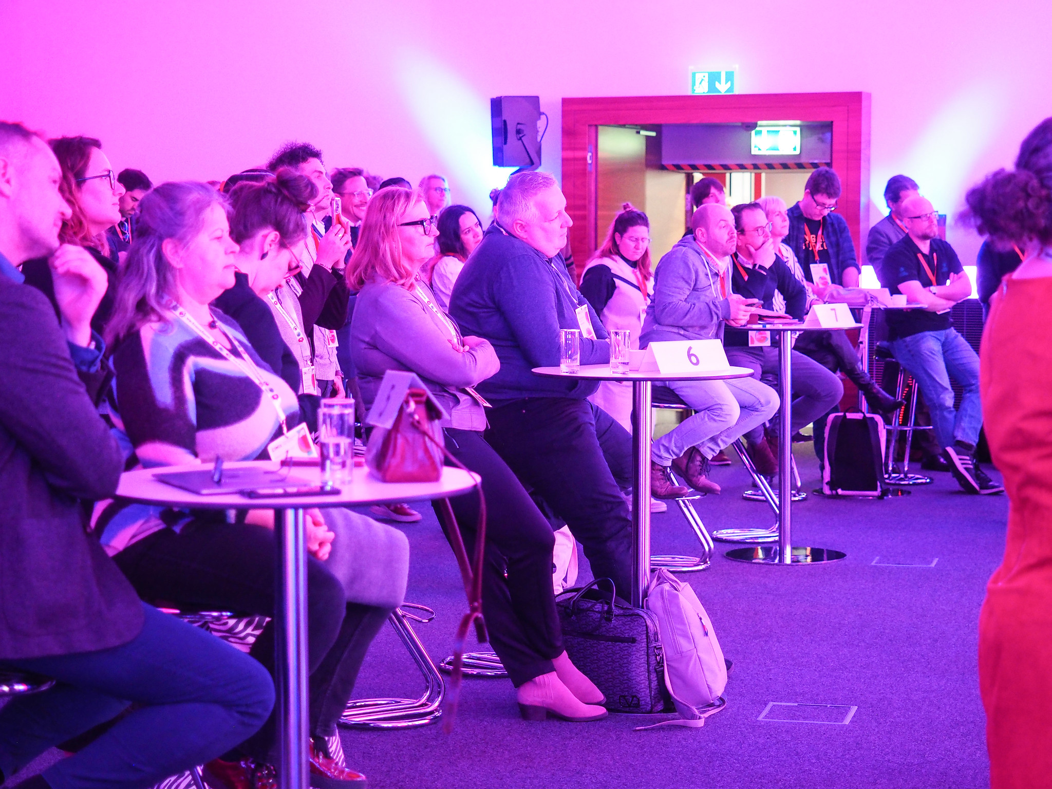 Eine Gruppe von Personen sitzt an runden Tischen und verfolgt aufmerksam eine Präsentation zum Thema "Connecting the dots 2026" in einem Konferenzraum mit rosa Beleuchtung. Die meisten schauen nach vorne, und einige haben Laptops oder Notizblöcke auf den Tischen liegen.