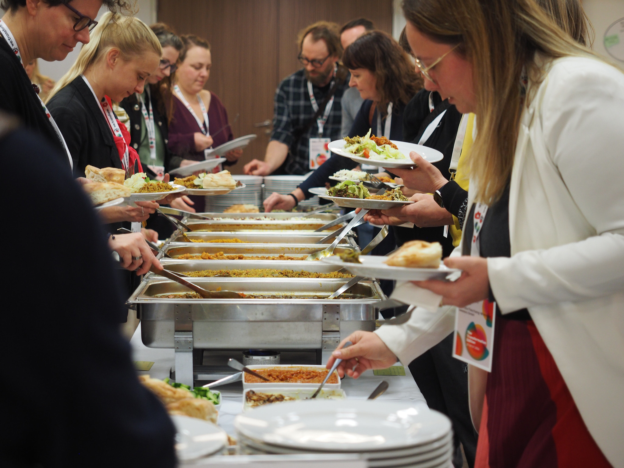 Auf dem CIMIx 2026 bedient sich eine Gruppe von Menschen an einem Buffet mit verschiedenen Gerichten, Salaten und Brot. Sie halten Teller und Utensilien in der Hand und stehen dicht beieinander in einem gut beleuchteten Innenraum.