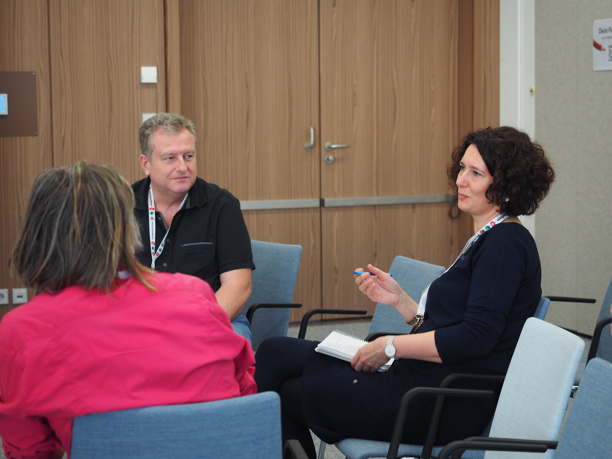 Auf der CIMIx 2026 sitzen drei Personen in einem Konferenzraum in einer kleinen Gruppe zusammen. Eine Frau hält einen Stift und ein Notizbuch in der Hand, während eine andere Person in einer rosa Jacke den Blick abwendet. Ein Mann hört aufmerksam zu, umgeben von leeren Stühlen.