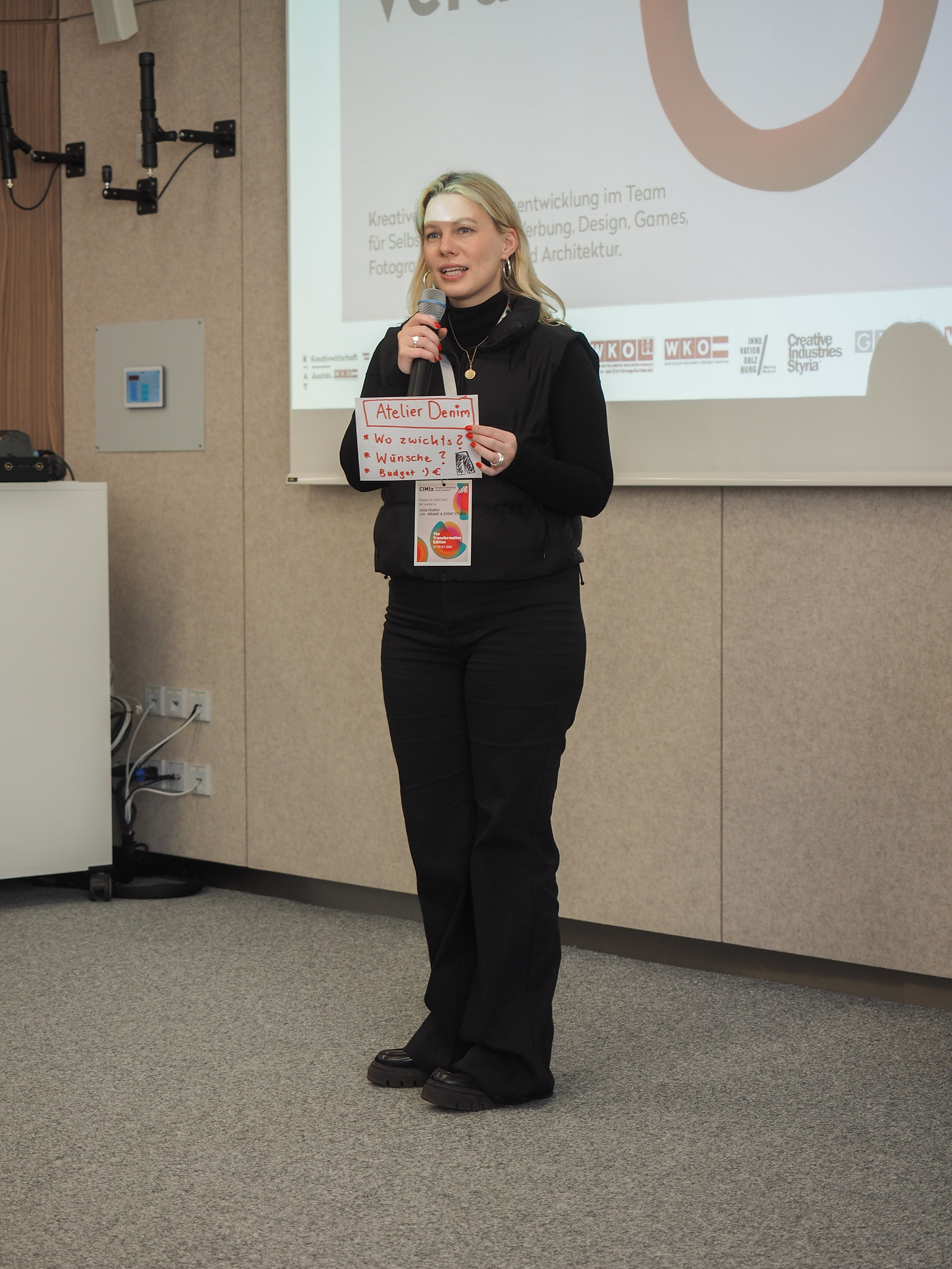 Eine schwarz gekleidete Frau steht und spricht vor einem projizierten Präsentationsbildschirm und hält ein kleines Poster mit der Aufschrift "Atelier Detail" auf der Veranstaltung CIMIx 2026. Der Raum scheint eine Konferenz oder ein Klassenzimmer zu sein.