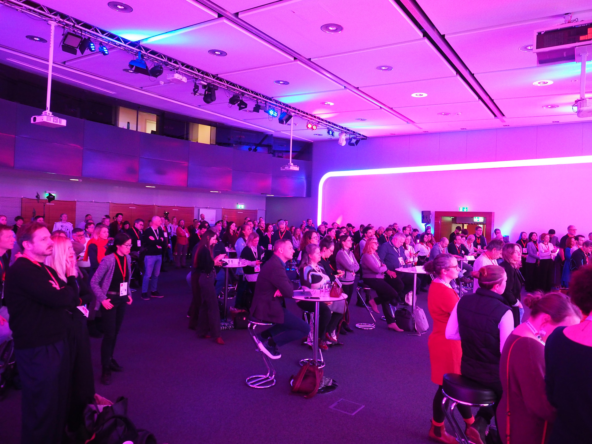 Eine große Gruppe von Menschen steht und sitzt an hohen Tischen in einem modernen, violett beleuchteten Konferenzraum und verfolgt aufmerksam eine "Connecting the dots 2026"-Präsentation. Einige tragen Namensschilder und Mäntel, und die Atmosphäre ist professionell und engagiert.