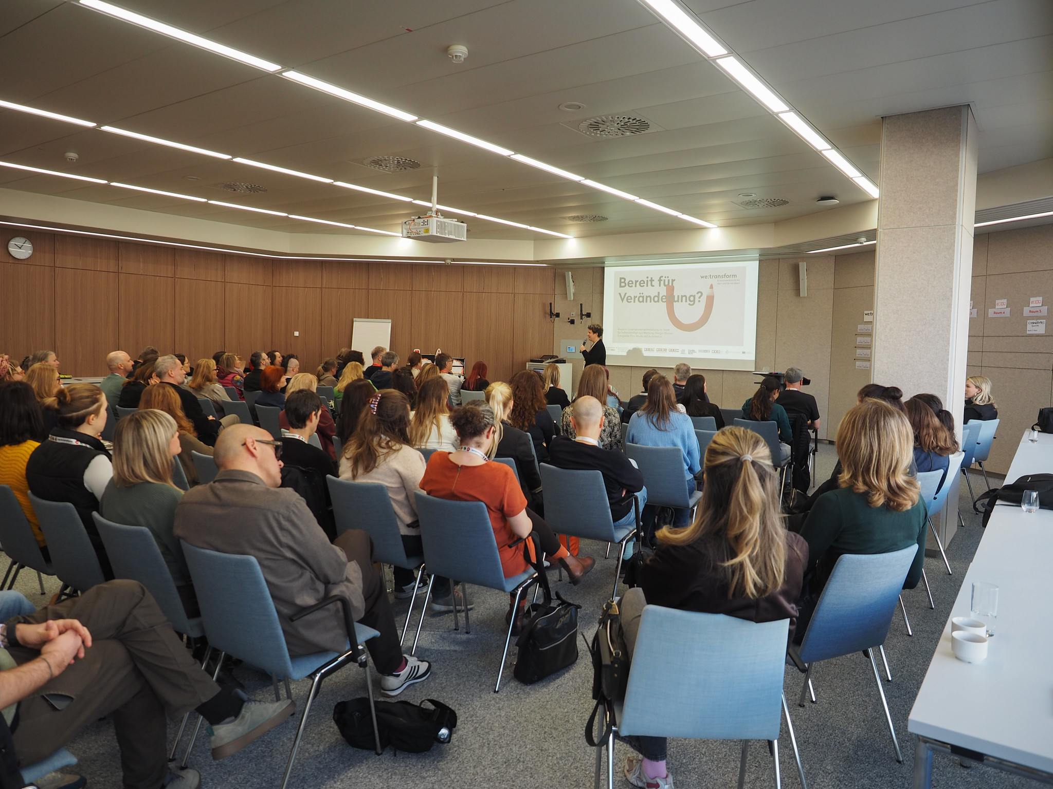 Eine Gruppe von Personen sitzt in einem Konferenzraum vor einem Präsentationsbildschirm mit der Aufschrift "Bereit für Veränderung?" und hört aufmerksam zu. Moderne Beleuchtung und Möbel bilden den Rahmen für die Veranstaltung, die Teil der Reihe "Connecting the Dots 2026" ist.