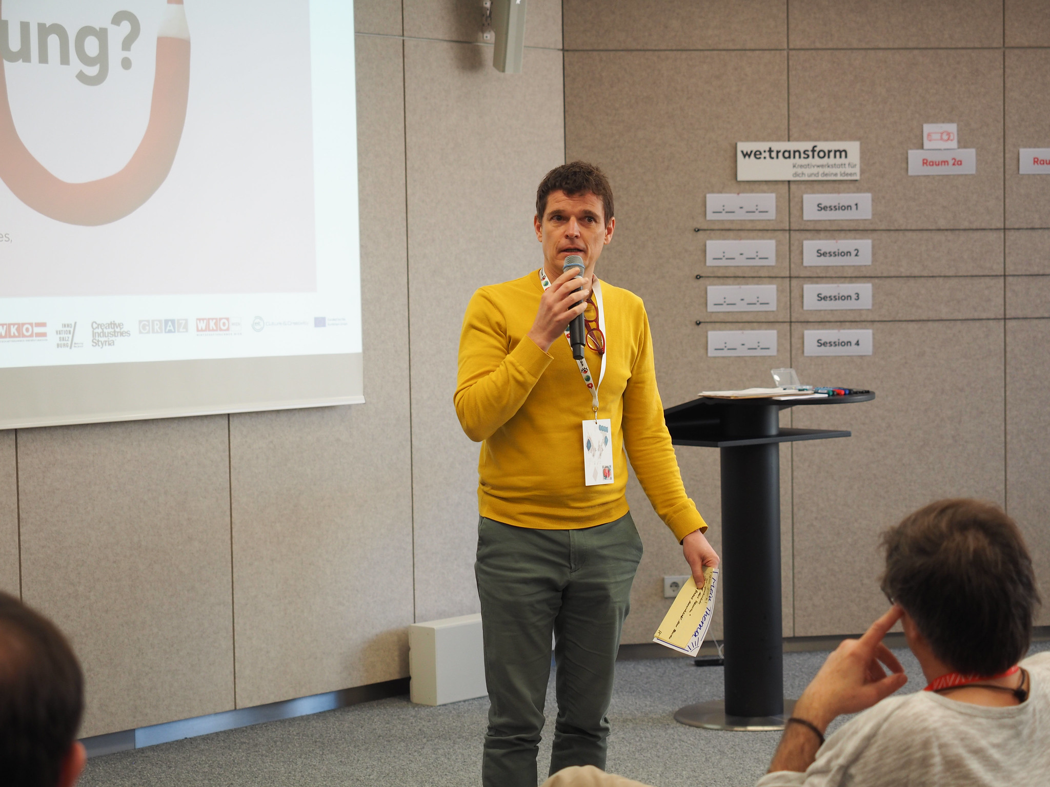 Ein Mann in einem gelben Pullover steht auf der Konferenz CIMIx 2026 und spricht in ein Mikrofon. Er hält eine Broschüre in der Hand und trägt ein Schlüsselband. Im Hintergrund sind eine Projektionsfläche und ein Podium zu sehen, teilweise sind auch Zuhörer zu erkennen.