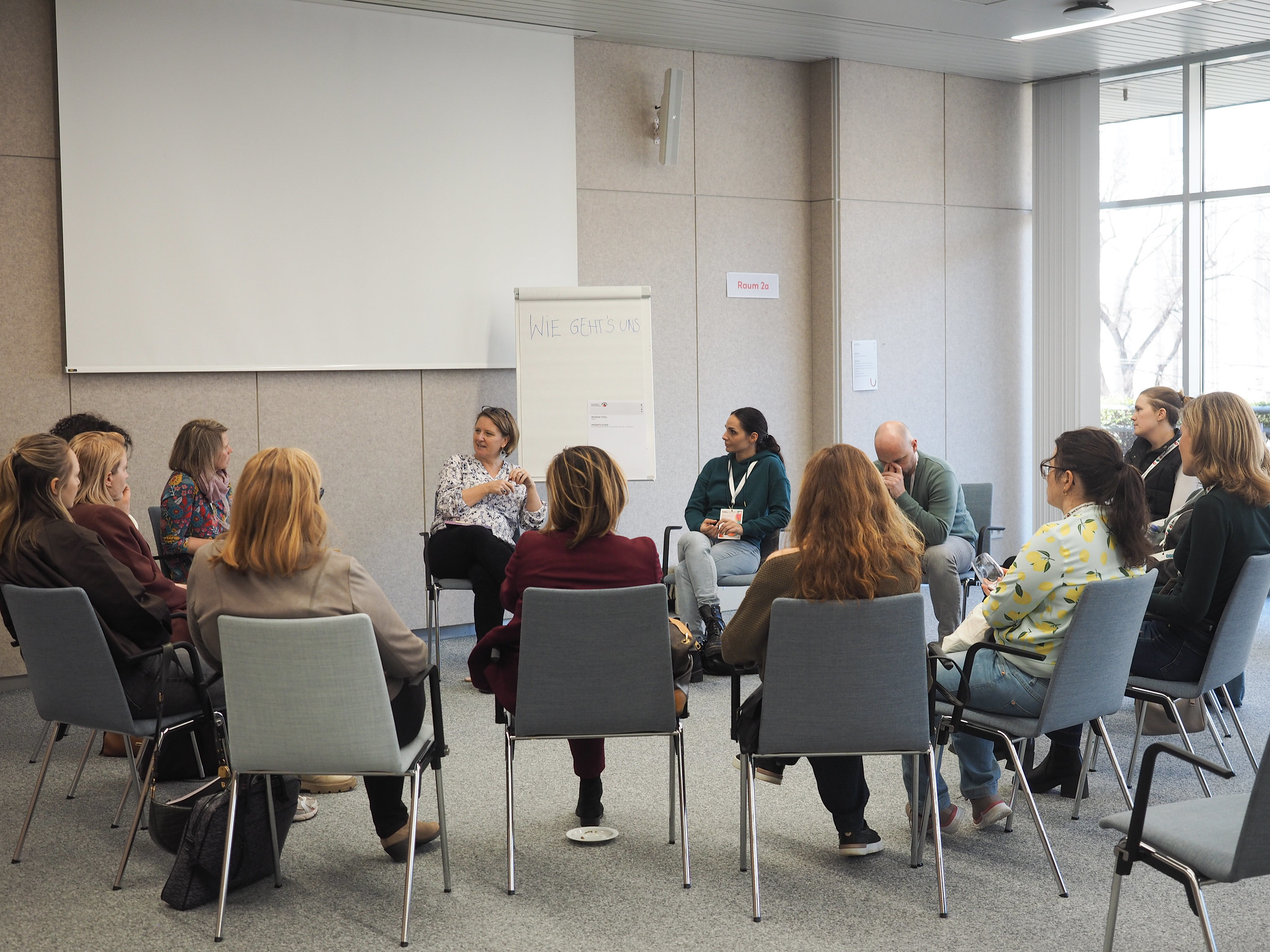 Eine Gruppe von Menschen sitzt in einem Stuhlkreis in einem modernen Konferenzraum des CIMIx 2026 und diskutiert. Eine Frau spricht, während die anderen aufmerksam zuhören. Im Hintergrund sind ein Flipchart und eine Projektionsfläche zu sehen.
