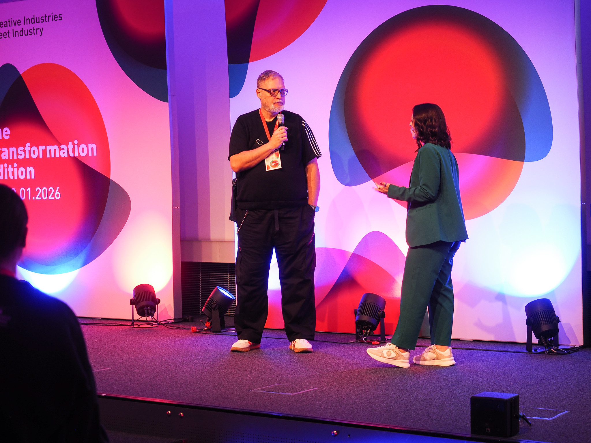 Ein Mann und eine Frau stehen auf der Bühne und sprechen in Mikrofone. Auf dem farbenfrohen Hintergrund sind kreisförmige Muster und Texte zu den Themen "Connecting the dots 2026", "Transformation" und "Innovation" zu sehen. Beide tragen legere Kleidung und sind in ein Gespräch vertieft.