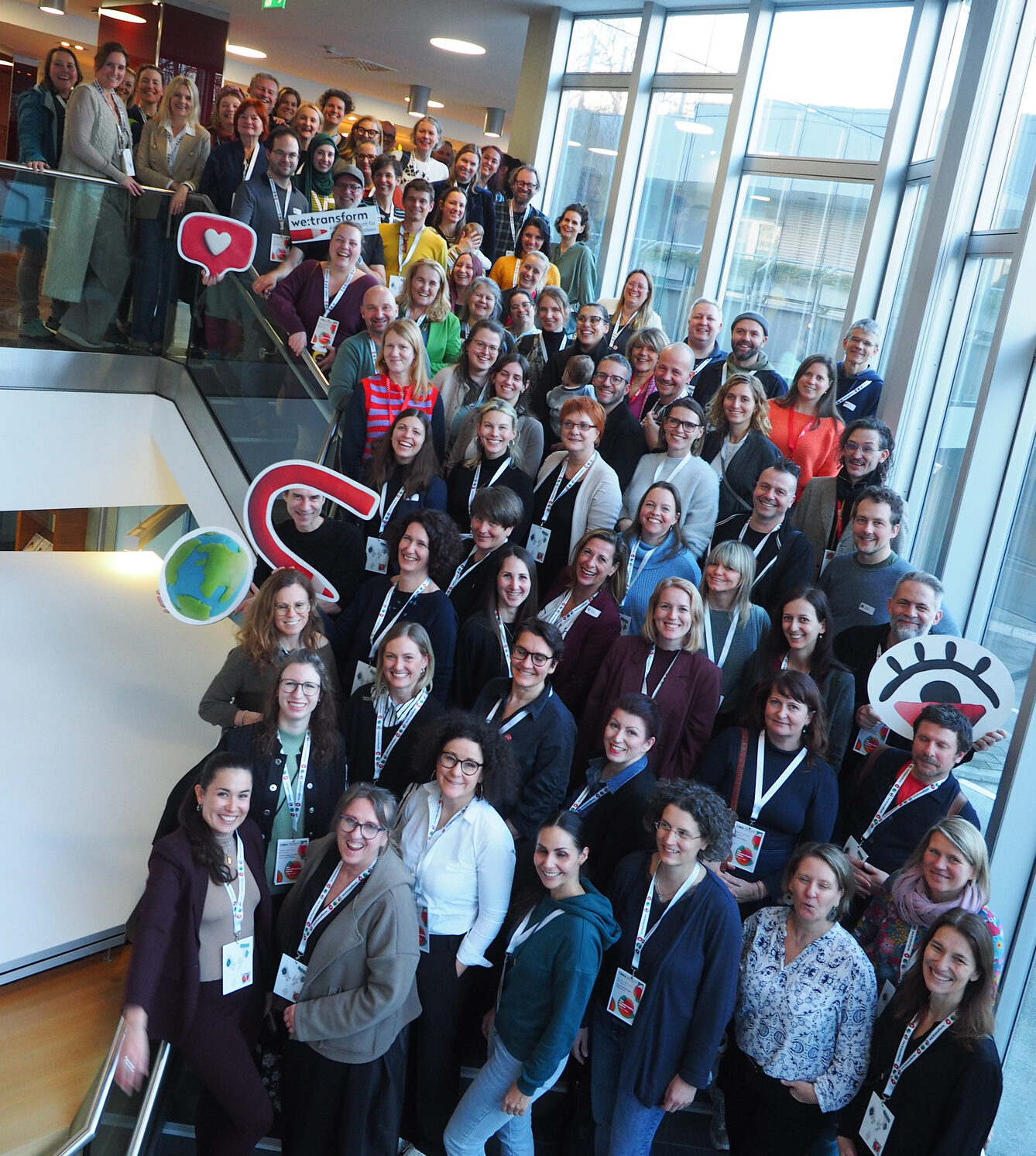 Eine große Gruppe von Menschen posiert gemeinsam auf einer Treppe in einem Innenraum, viele lächeln in die Kamera. Einige halten bunte Schilder mit Symbolen und CIMIx 2026-Logos in der Hand, und alle tragen Namensschilder. Große Fenster lassen natürliches Licht hinter ihnen herein.