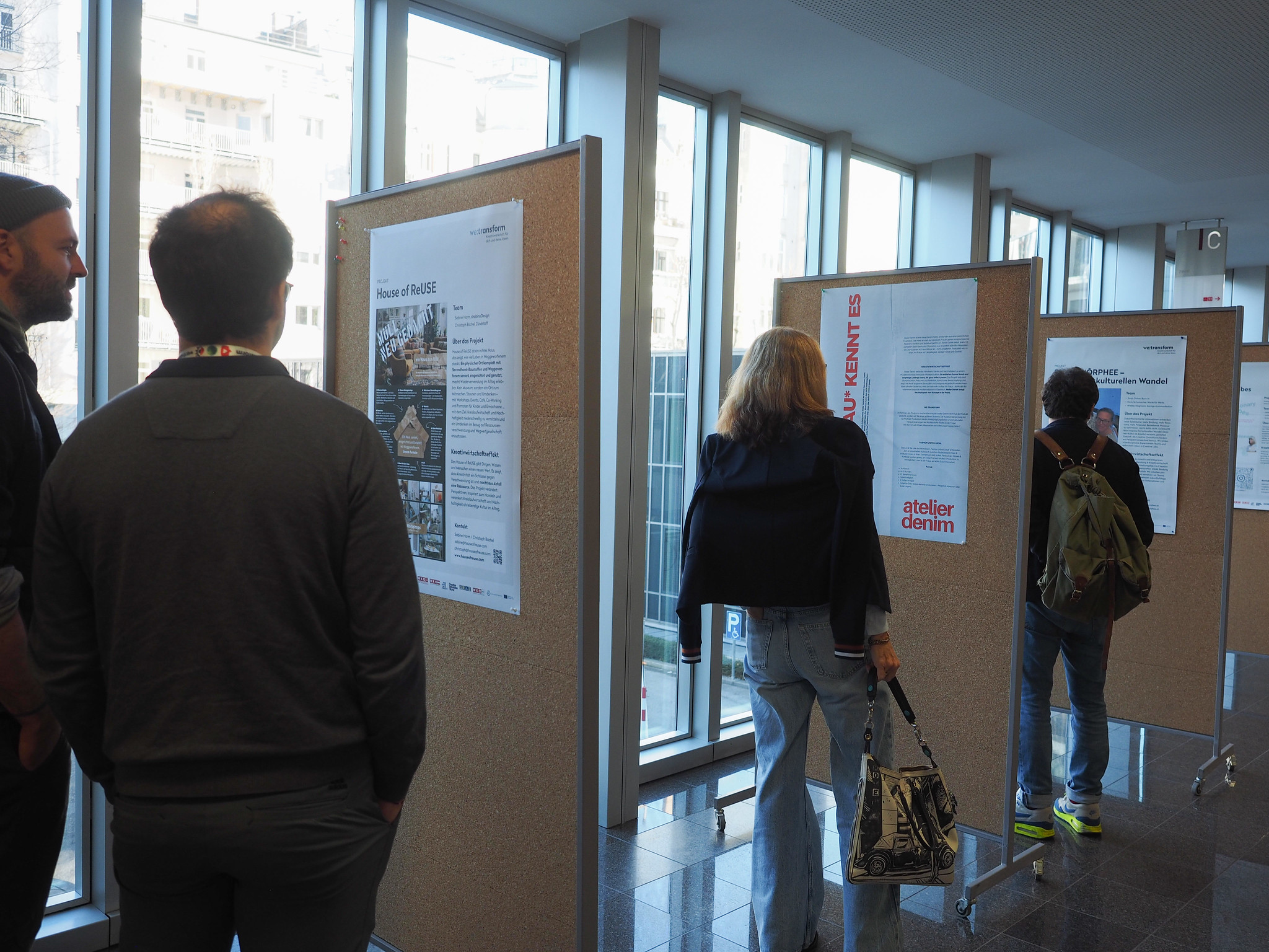 Mehrere Personen stehen in einem hell erleuchteten Flur des CIMIx 2026 und lesen die auf Korkplatten angebrachten Poster. Die Poster enthalten Texte und Bilder, während große Fenster im Hintergrund natürliches Licht hereinlassen.