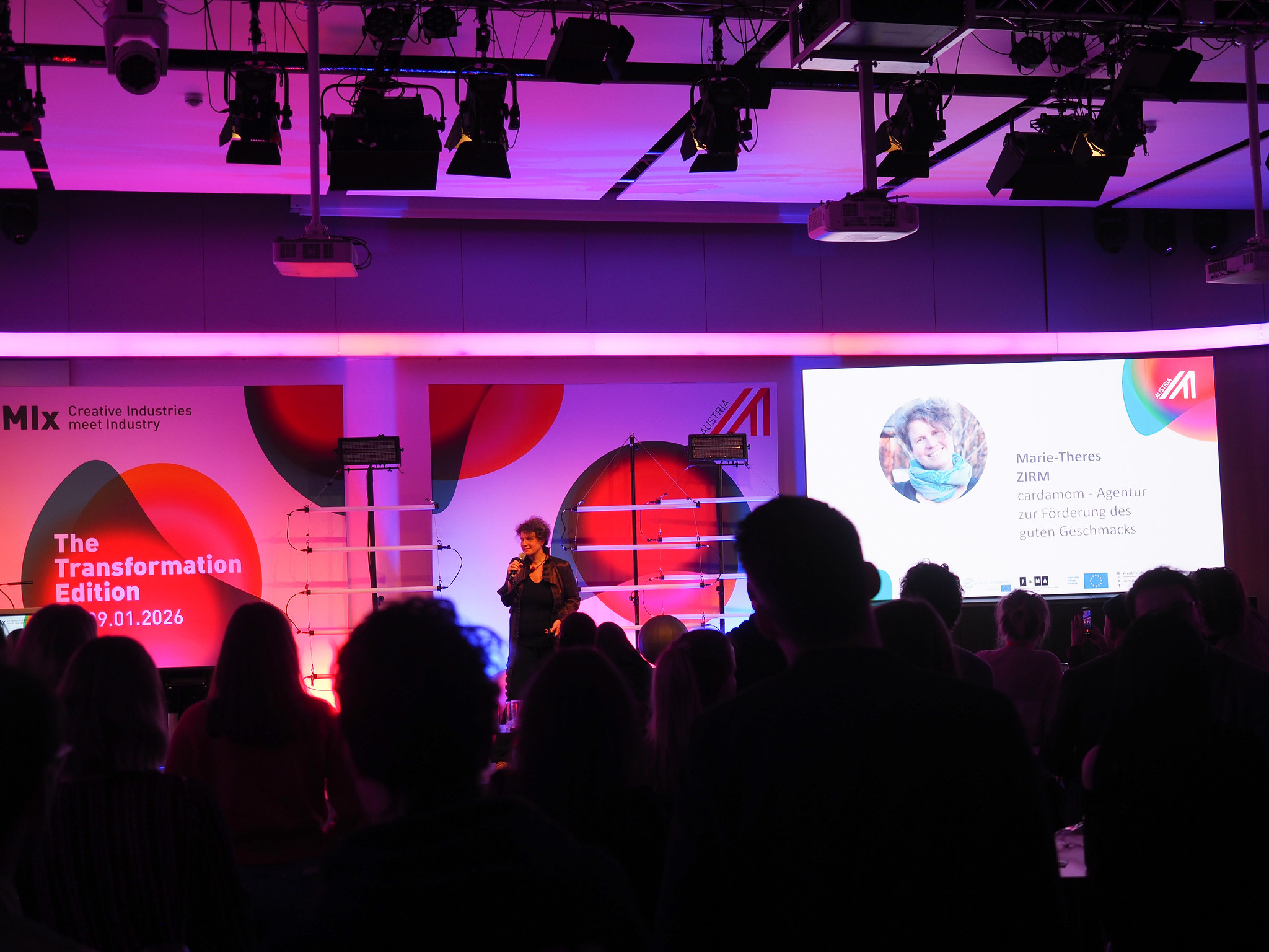 Ein Redner steht auf der Bühne und wendet sich an das Publikum einer Veranstaltung. Auf einem großen Bildschirm werden das Kopfbild von Marie-Theres Zirm und Informationen neben bunten Grafiken und dem Text The Transformation Edition, Connecting the dots 2026, 29.01.2026 angezeigt.