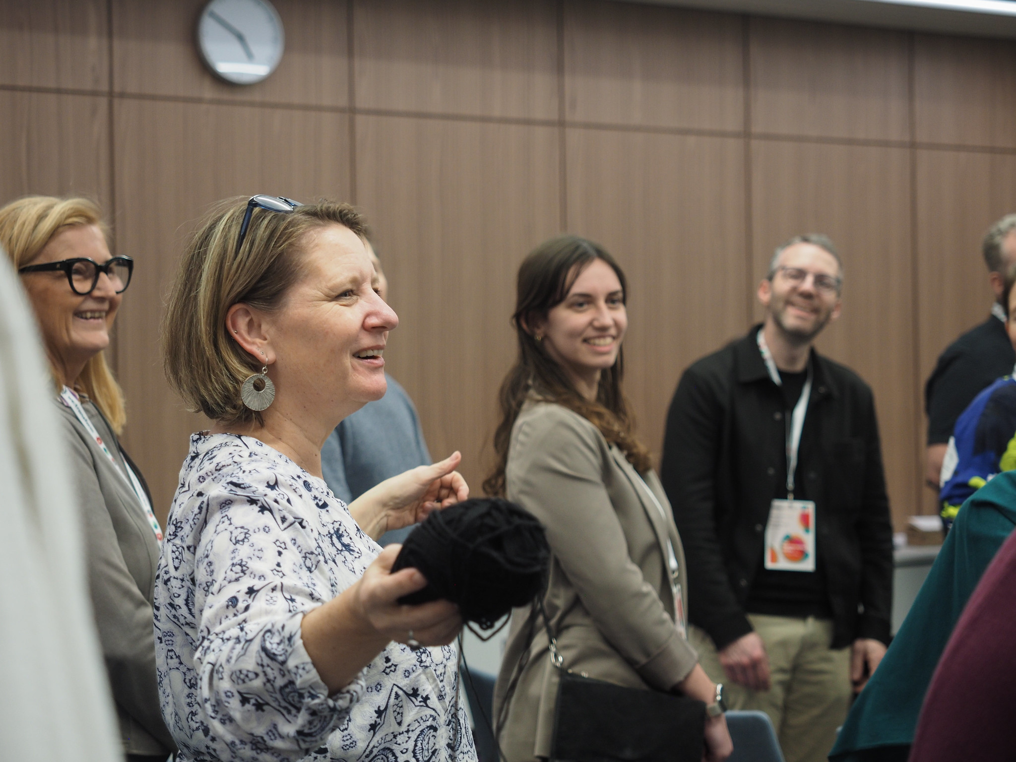 Eine Gruppe Erwachsener steht in einem Raum des CIMIx 2026, lächelt und unterhält sich. Eine Frau im Vordergrund, die ein Garnknäuel in der Hand hält, scheint zu sprechen, während die anderen um sie herum zuhören und lachen. An der Wand hinter ihnen ist eine Uhr zu sehen.