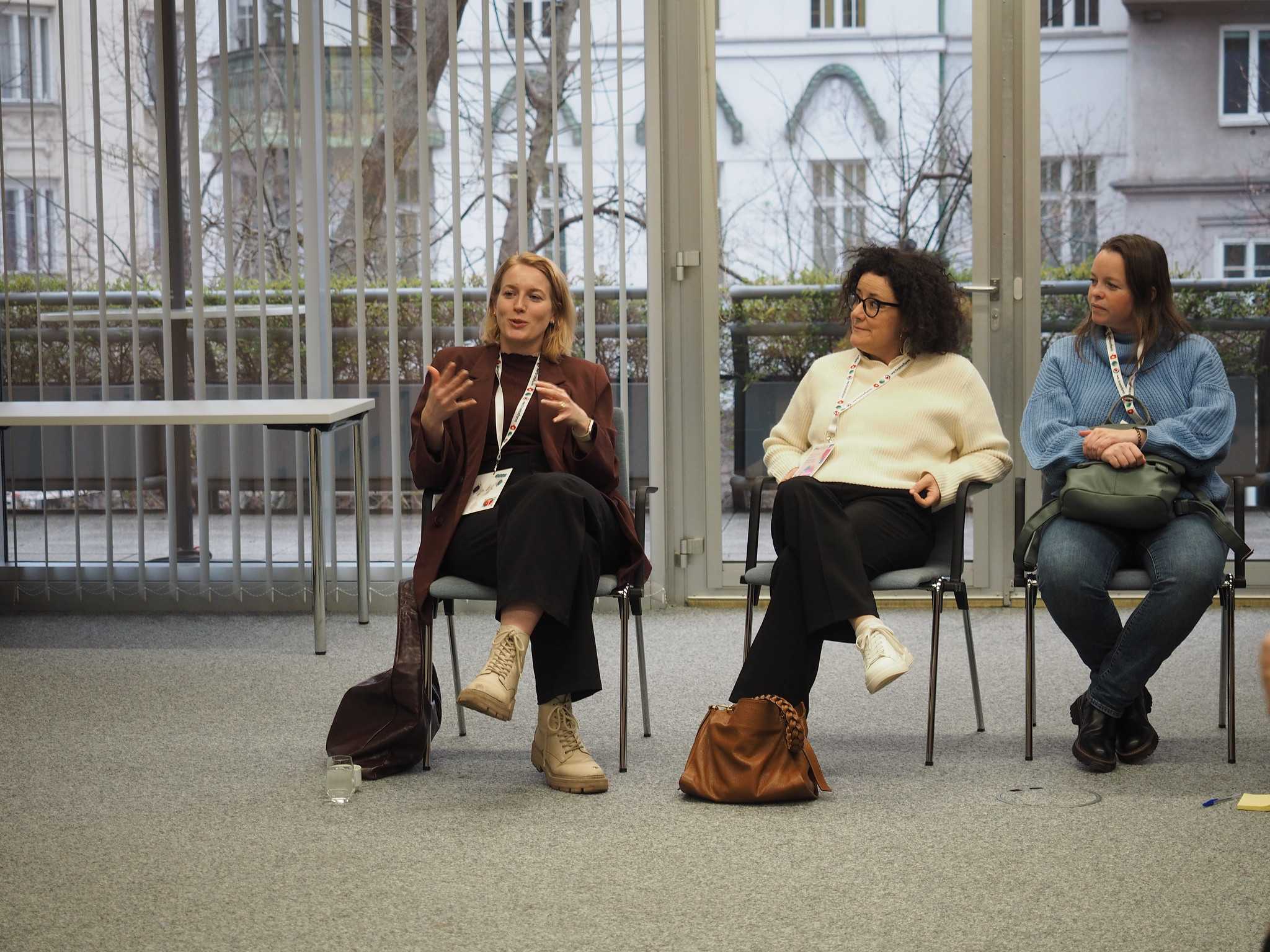 Drei Frauen sitzen in einem Raum auf Stühlen und diskutieren über CIMIx 2026. Eine Frau gestikuliert, während sie spricht, und die beiden anderen hören aufmerksam zu. Große Fenster im Hintergrund zeigen ein Gebäude und blattlose Bäume draußen.