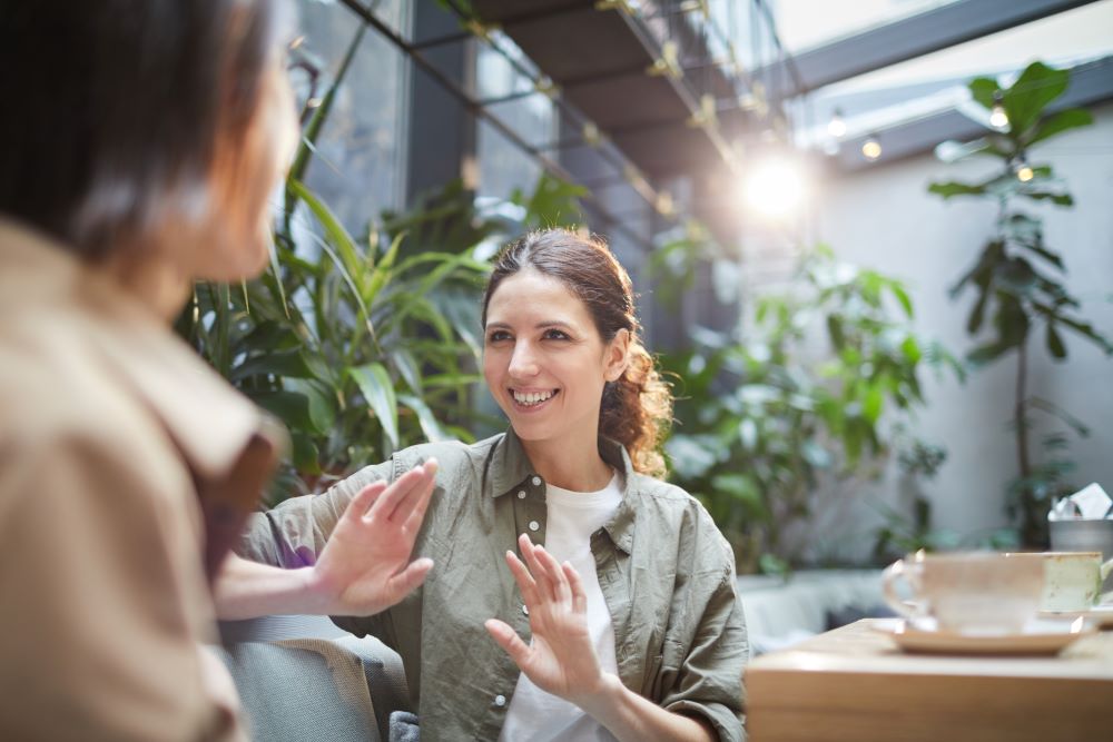 Eine Frau l&auml;chelt und gestikuliert mit den H&auml;nden, w&auml;hrend sie sich mit einer anderen Person in einem hellen, modernen Caf&eacute; mit Gr&uuml;npflanzen unterh&auml;lt. Eine Tasse und eine Untertasse stehen vor ihr auf dem Tisch.