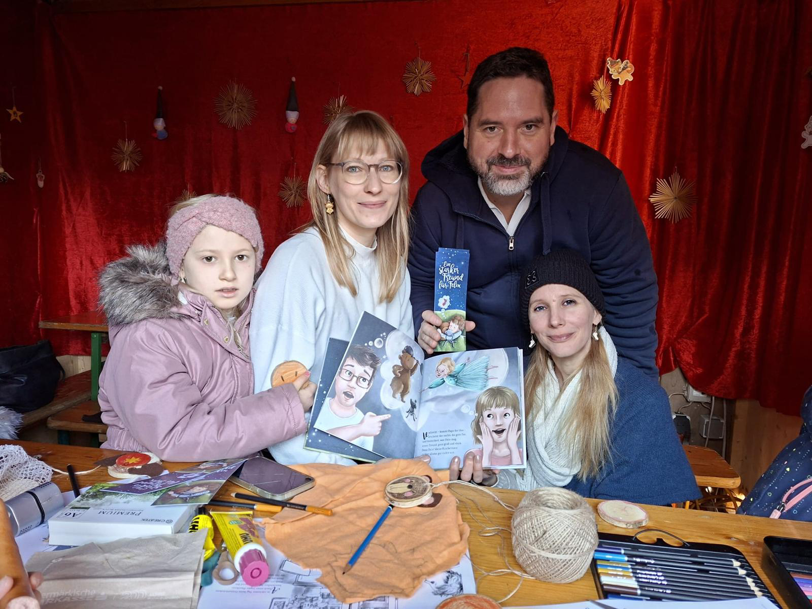 Vier Personen - zwei Erwachsene und zwei Kinder - stehen in einem Raum vor einem Tisch mit Malutensilien und zeigen ein illustriertes Buch. Der Hintergrund ist rot mit hängenden Dekorationen. Alle lächeln und scheinen glücklich zu sein.