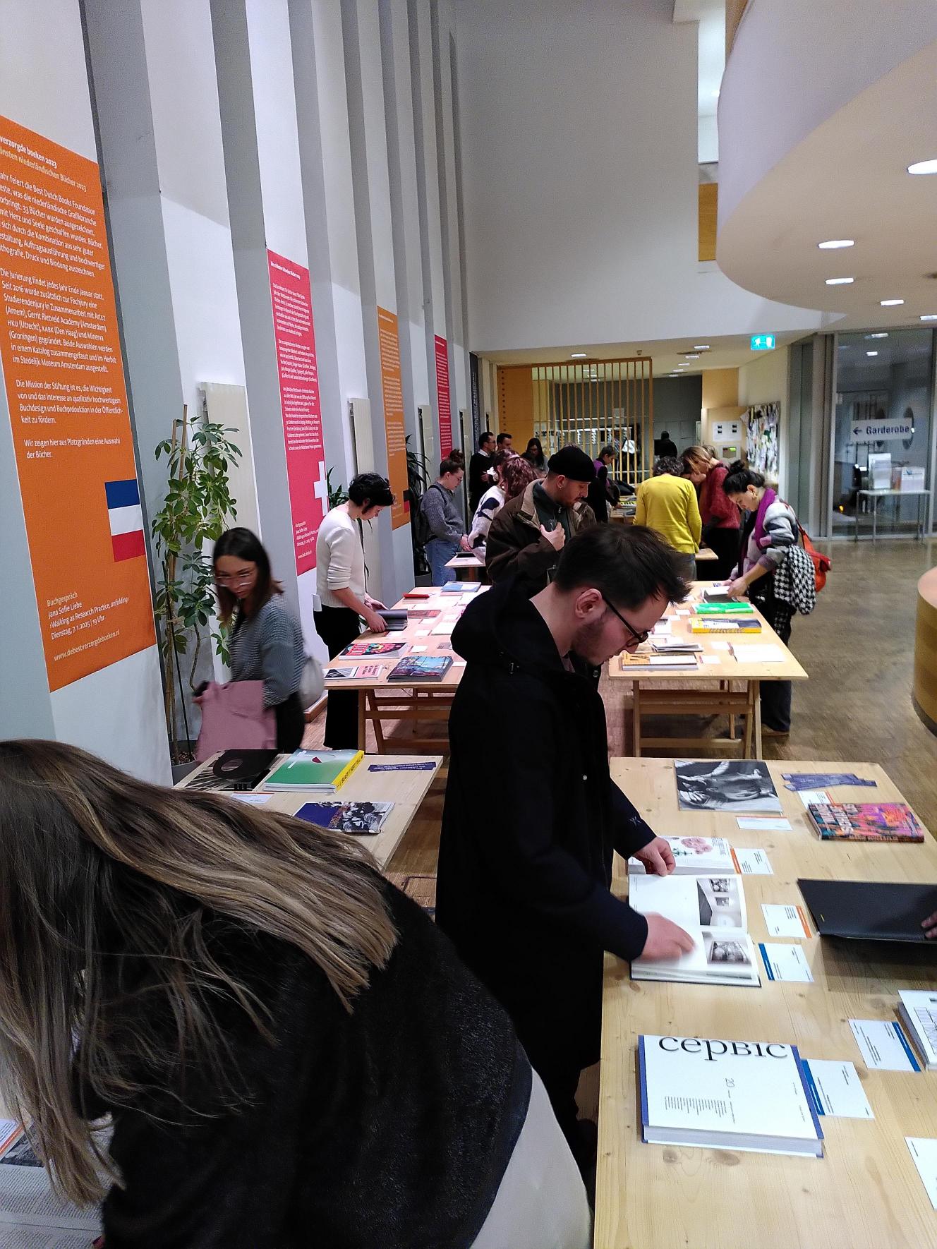 In einem hellen, modernen Innenraum der Ars Electronica mit orangefarbenen Informationsplakaten an den W&auml;nden und gro&szlig;en Fenstern stehen Menschen an langen Tischen und bl&auml;ttern in B&uuml;chern und Zeitschriften.