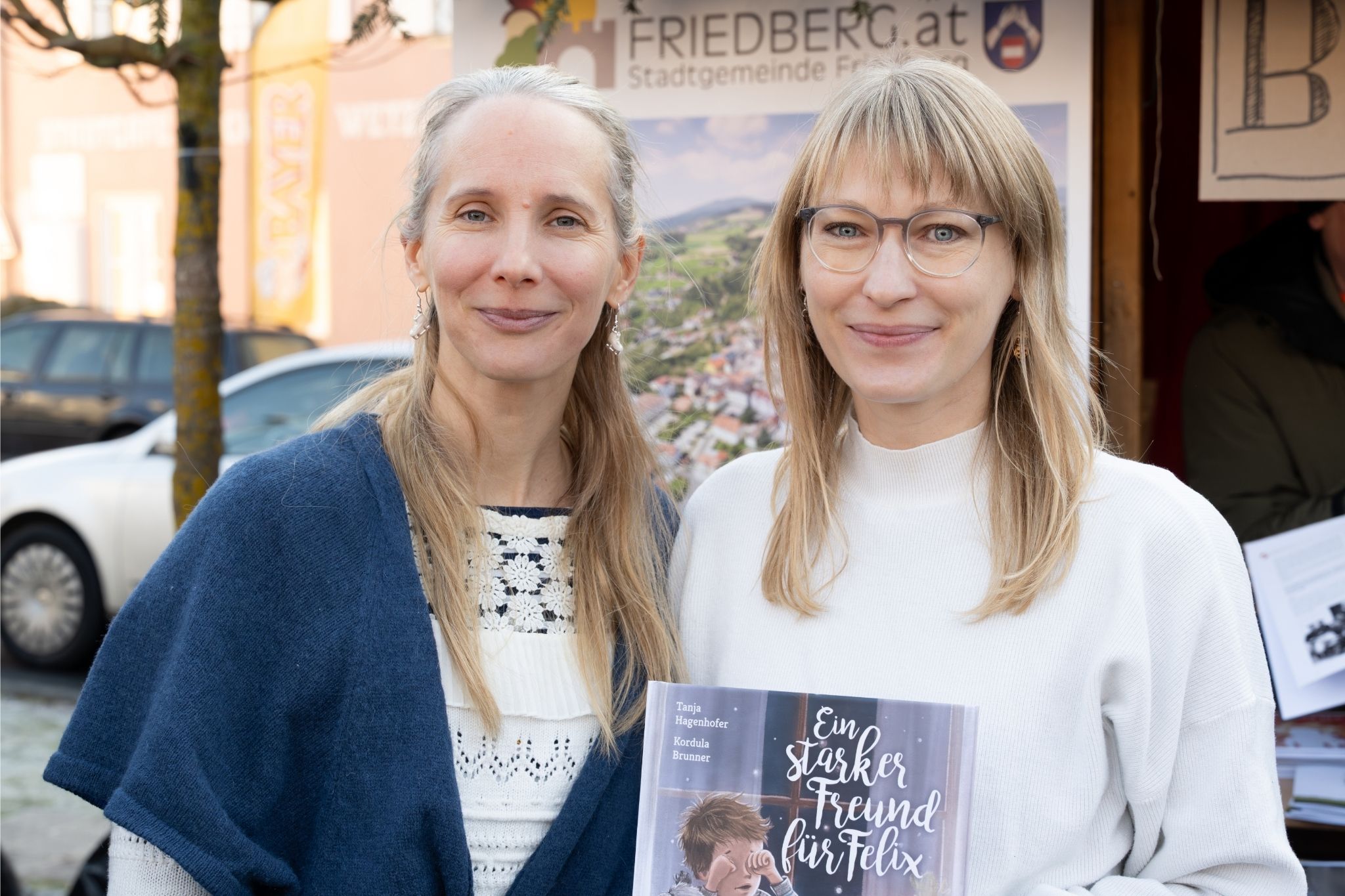 Zwei Frauen stehen dicht beieinander und lächeln in die Kamera im Freien. Eine Frau hält ein Buch mit dem Titel Ein starker Freund für Felix in der Hand. Im Hintergrund sind ein Auto, Gebäude und ein Schild mit der Aufschrift FRIEDBERG.at zu sehen.