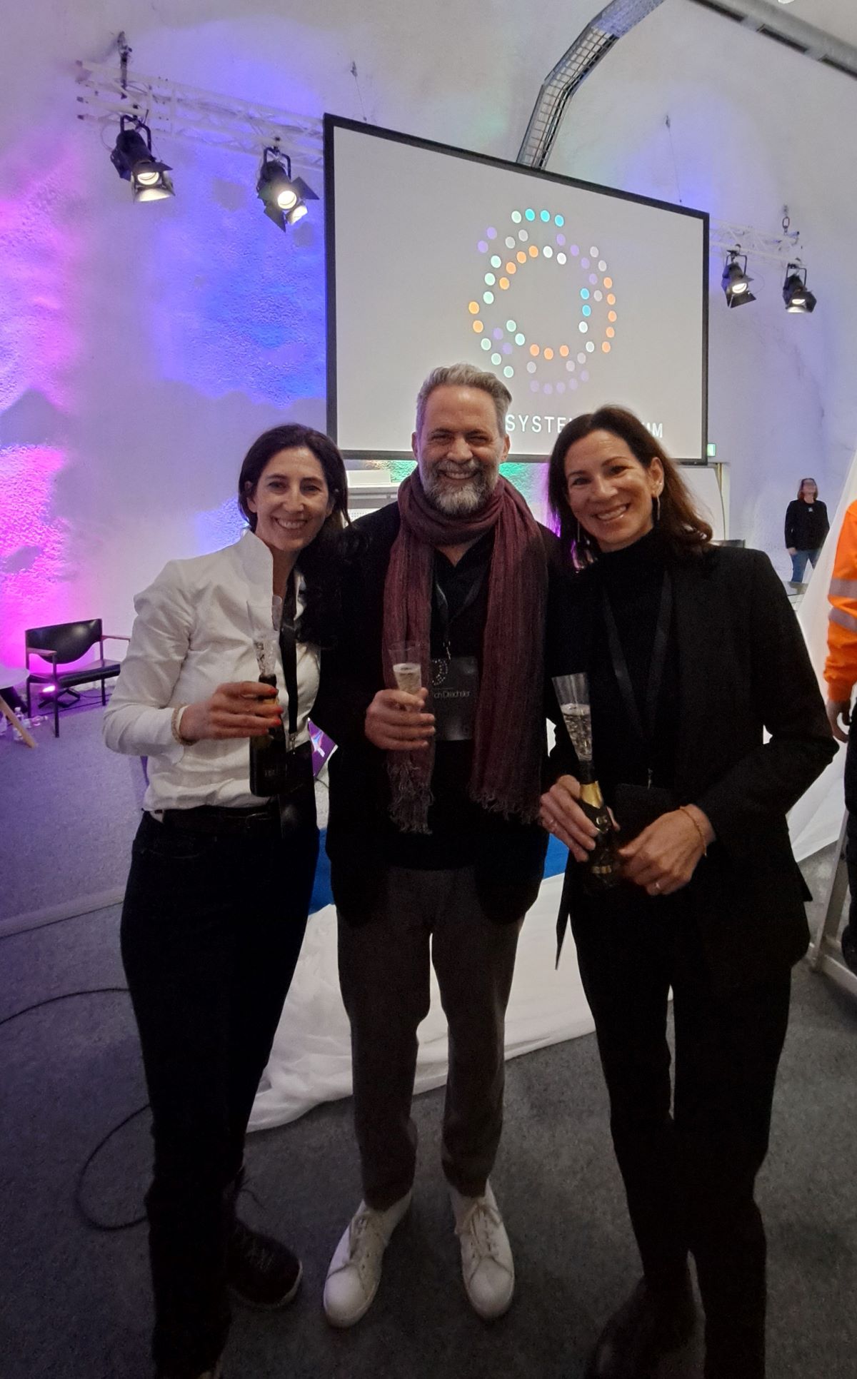 Drei Personen, darunter der Musiker Ulrich Drechsler, stehen in einem Innenraum zusammen, lächeln und halten Gläser. Hinter ihnen ist ein großer Bildschirm mit einem kreisförmigen Logo aus farbigen Punkten zu sehen, im Hintergrund sind Bühnenbeleuchtung und Veranstaltungstechnik zu sehen.