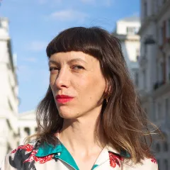 Eine Frau mit schulterlangem braunem Haar und stumpfem Pony steht in einer Stadt im Freien, trägt eine geblümte Bluse und roten Lippenstift und blickt selbstbewusst in die Kamera, während im Hintergrund Gebäude und ein blauer Himmel zu sehen sind.