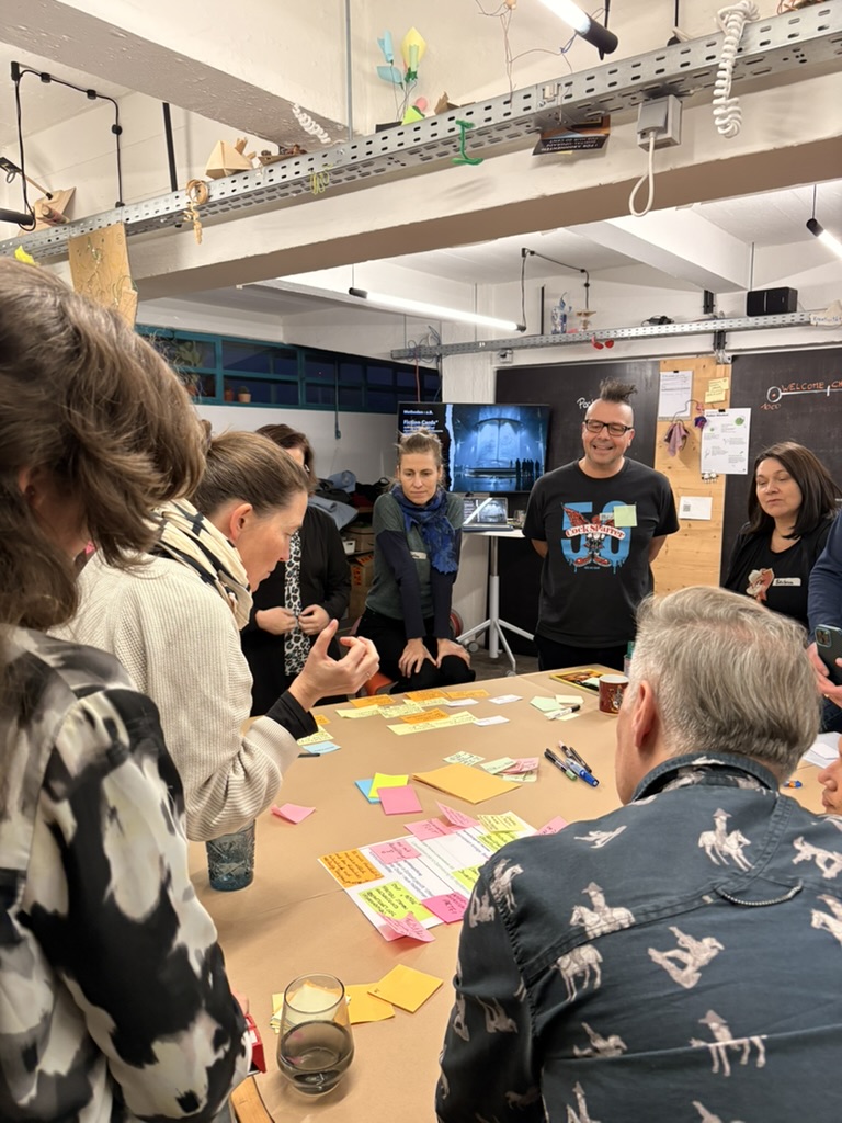 Eine Gruppe von Menschen versammelt sich um einen Tisch, der mit bunten Haftnotizen und Papieren bedeckt ist. Sie diskutieren und arbeiten aktiv in einem kreativen Methoden BarCamp Arbeitsbereich zusammen. Im Hintergrund sind Regale und Notizen zu sehen.