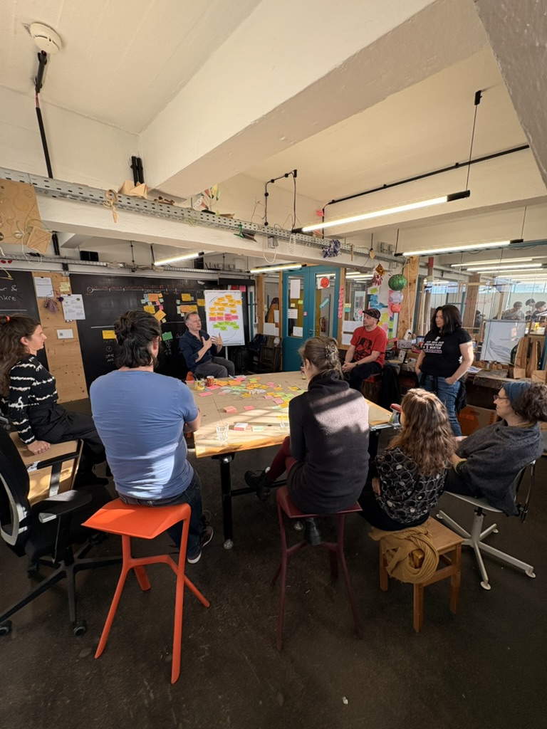 Eine Gruppe von Menschen sitzt um einen großen Tisch mit Papieren und Haftnotizen in einem hell erleuchteten, kreativen Arbeitsbereich und beteiligt sich aktiv an einer Diskussion oder einem Workshop im Rahmen eines Methoden-Barcamps.