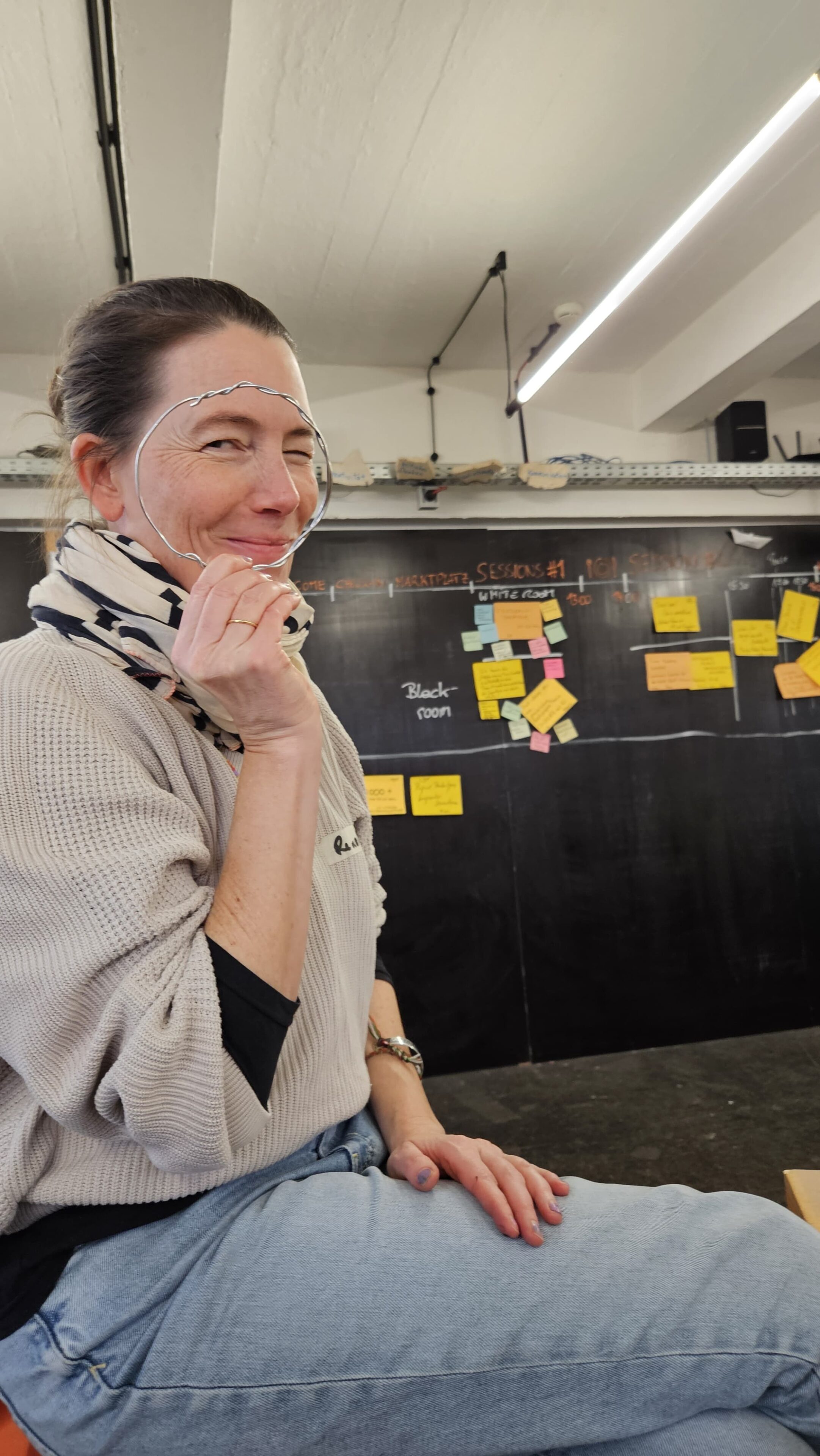 Eine Frau mit Kopftuch hält sich einen Draht vor das Gesicht und kneift spielerisch ein Auge zu. Hinter ihr füllen bunte Klebezettel die Tafel - eine lebendige Methoden-BarCamp-Szene in einem kreativen Klassenzimmer.