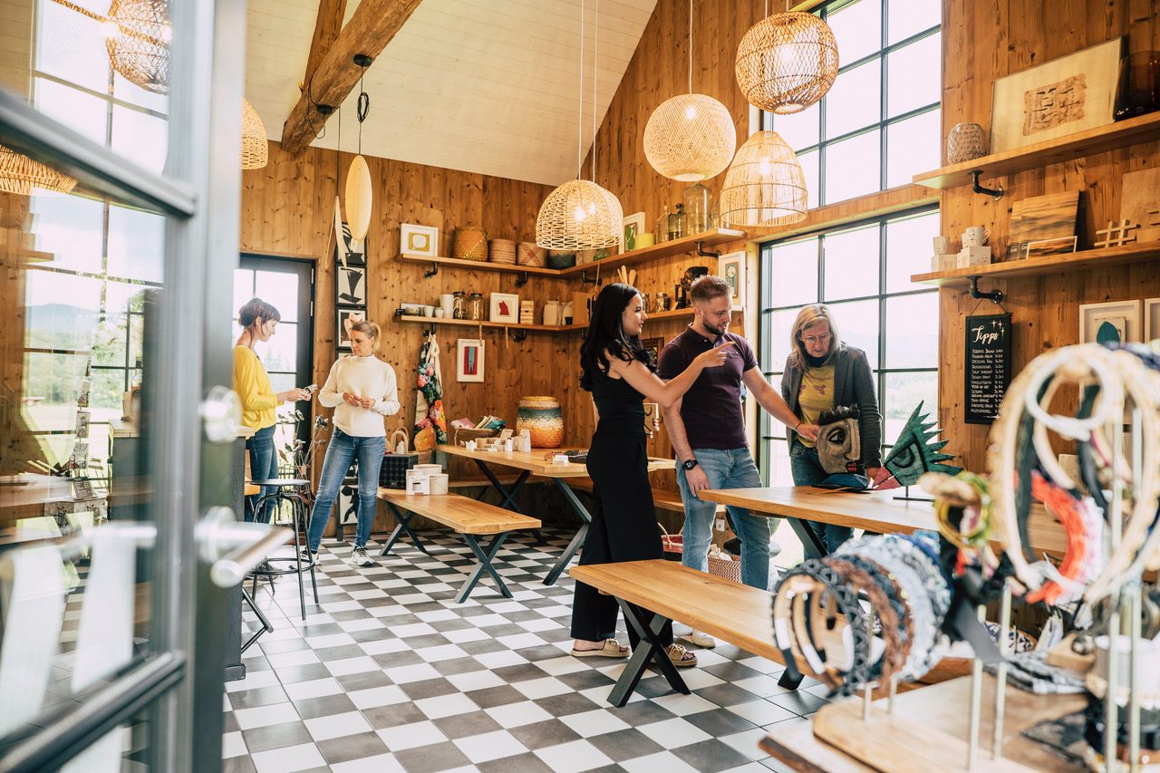 Eine Gruppe von fünf Personen trifft sich im Design ab Hof, einem gemütlichen, sonnendurchfluteten Laden mit Holzwänden, großen Fenstern und karierten Böden, und stöbert in handgefertigten Produkten. Handgefertigtes Kunsthandwerk und Schmuck sind auf Tischen und in Regalen ausgestellt. Rufen Sie 2026 für weitere Informationen an.