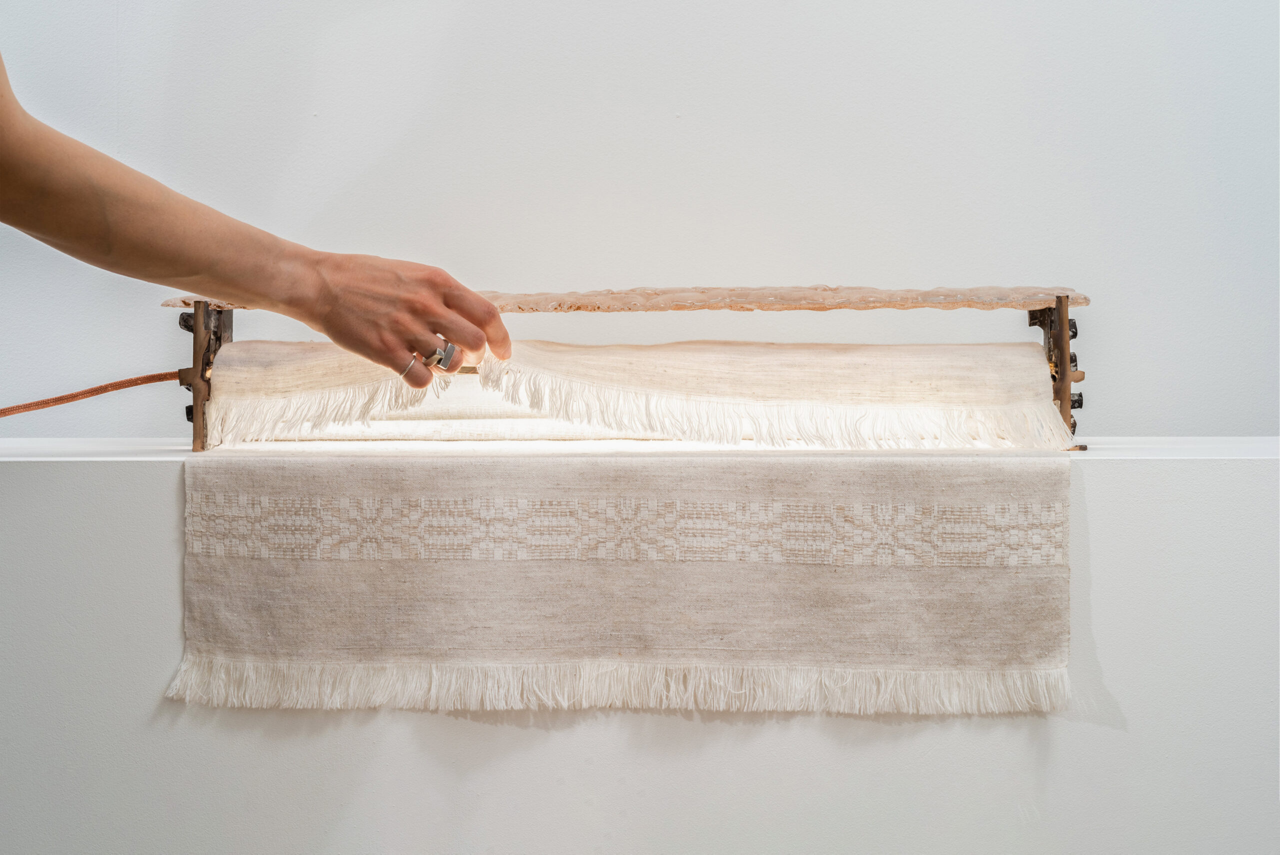 A hand - perhaps that of a master or apprentice - lifts a piece of woven fabric with frayed edges from a small wooden loom attached to a white wall. The fabric has a subtle decorative pattern.