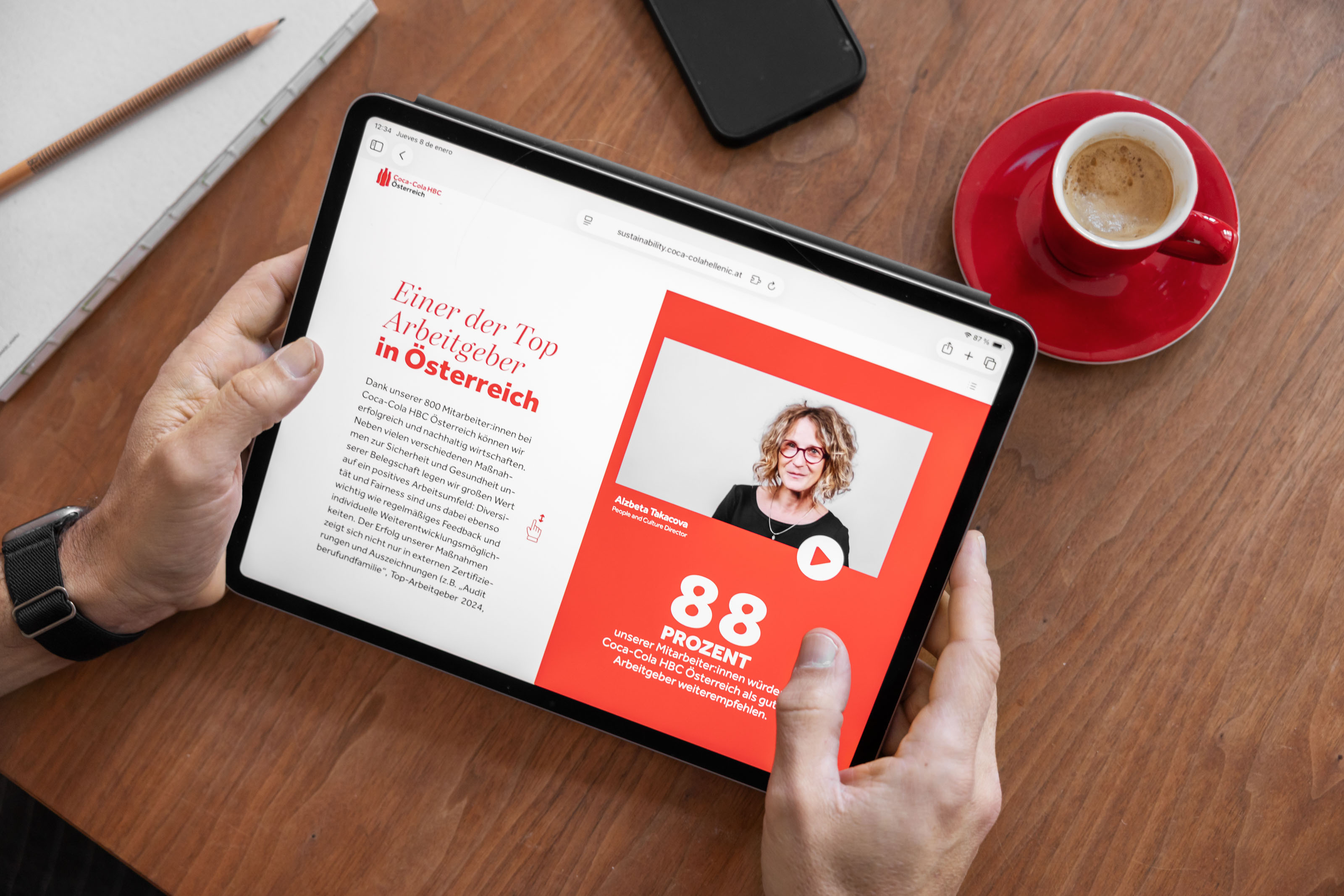 Eine Person hält ein Tablet in der Hand, auf dem eine deutschsprachige Website über einen Top-Arbeitgeber in Österreich angezeigt wird. Darauf sind das Porträt einer Frau, eine Play-Taste und die Zahl 88 Prozent auf rotem Hintergrund zu sehen. Eine Tasse Kaffee steht in der Nähe auf einem Holztisch.