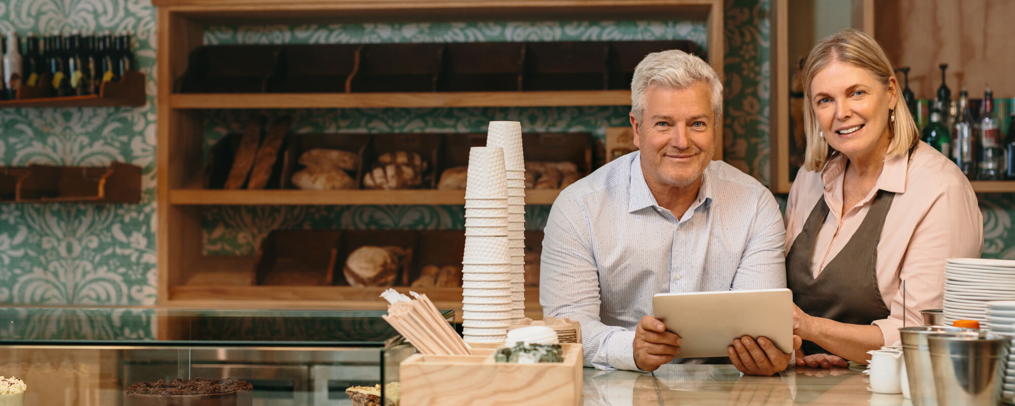 Ein Mann und eine Frau stehen hinter dem Tresen einer Bäckerei oder eines Cafés und lächeln in die Kamera. Der Mann hält ein Tablet in der Hand, und in der Auslage stehen Kaffeetassen, Gebäck und Brot. Im Hintergrund befinden sich Regale und gemusterte Tapeten.
