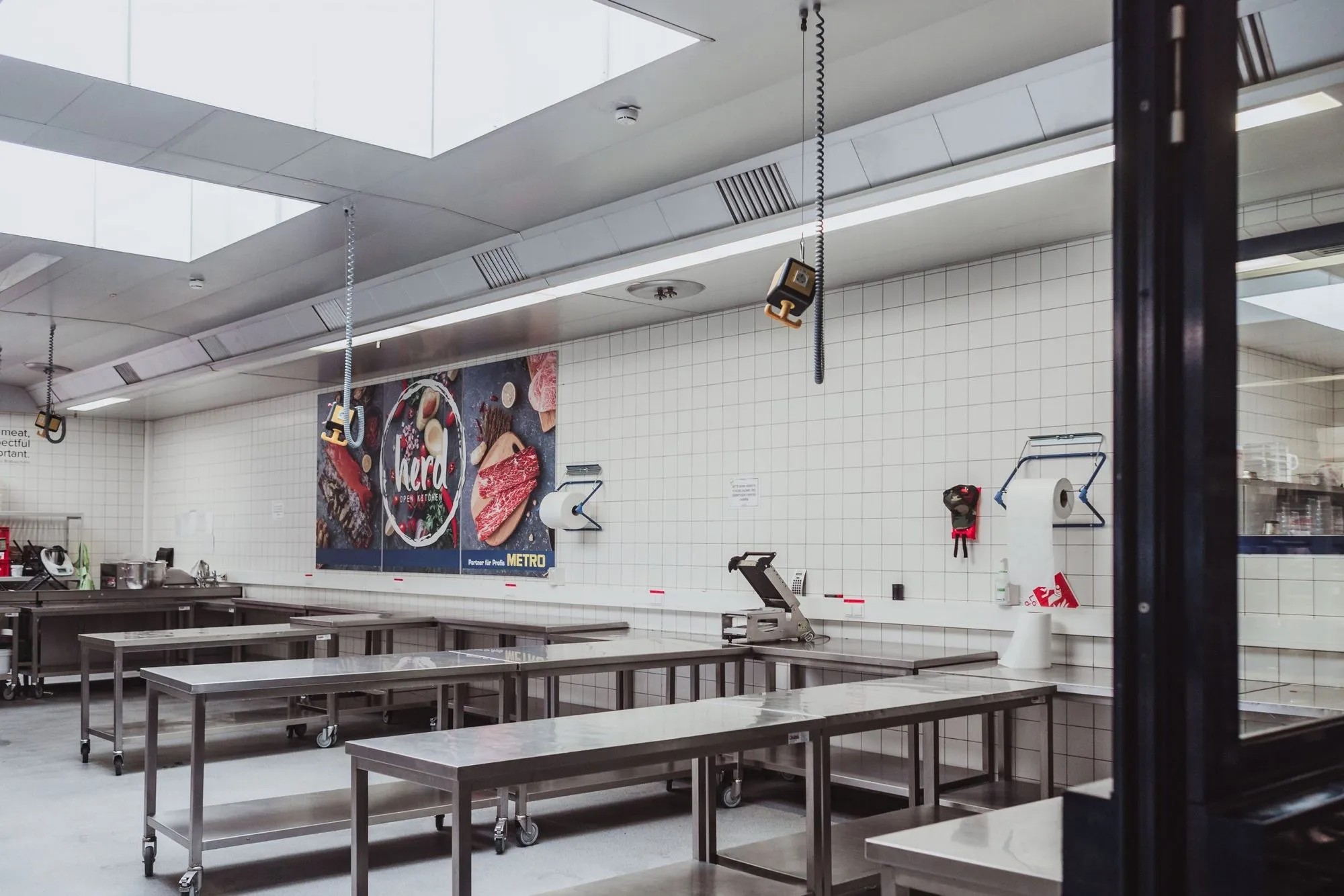 Eine saubere, leere Großküche mit Arbeitstischen aus rostfreiem Stahl, gefliesten Wänden, Oberlichtern und Geräten zur Lebensmittelzubereitung; an der Rückwand hängt ein großes Lebensmittelposter.