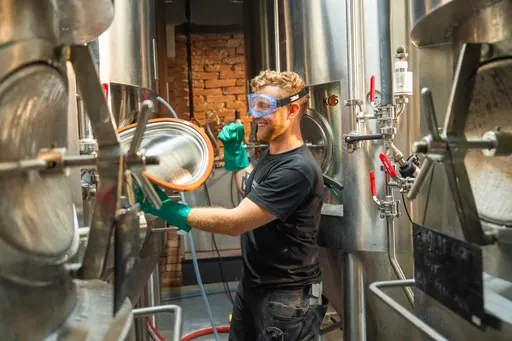 Ein Mann mit Schutzbrille und grünen Handschuhen reinigt mit einem Schlauch das Innere eines großen Brauerei-Tanks aus Edelstahl in einer industriellen Umgebung.