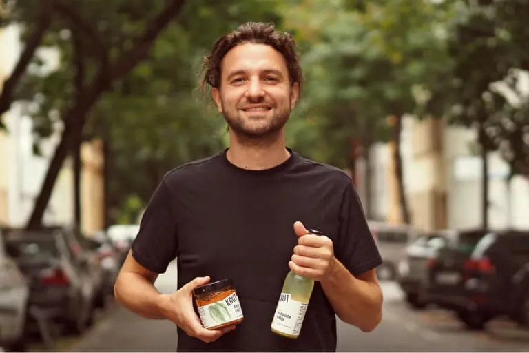 Ein lächelnder Mann mit langen Haaren in einem Pferdeschwanz und einem schwarzen Hemd steht auf einer von Bäumen gesäumten Straße und hält in der einen Hand ein Glas und in der anderen eine Flasche. Auf beiden Seiten der Straße sind Autos geparkt.