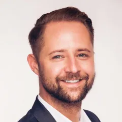 Ein Mann mit kurzen braunen Haaren und einem Bart lächelt, während er eine dunkle Anzugsjacke und ein weißes Hemd vor einem schlichten hellen Hintergrund trägt.