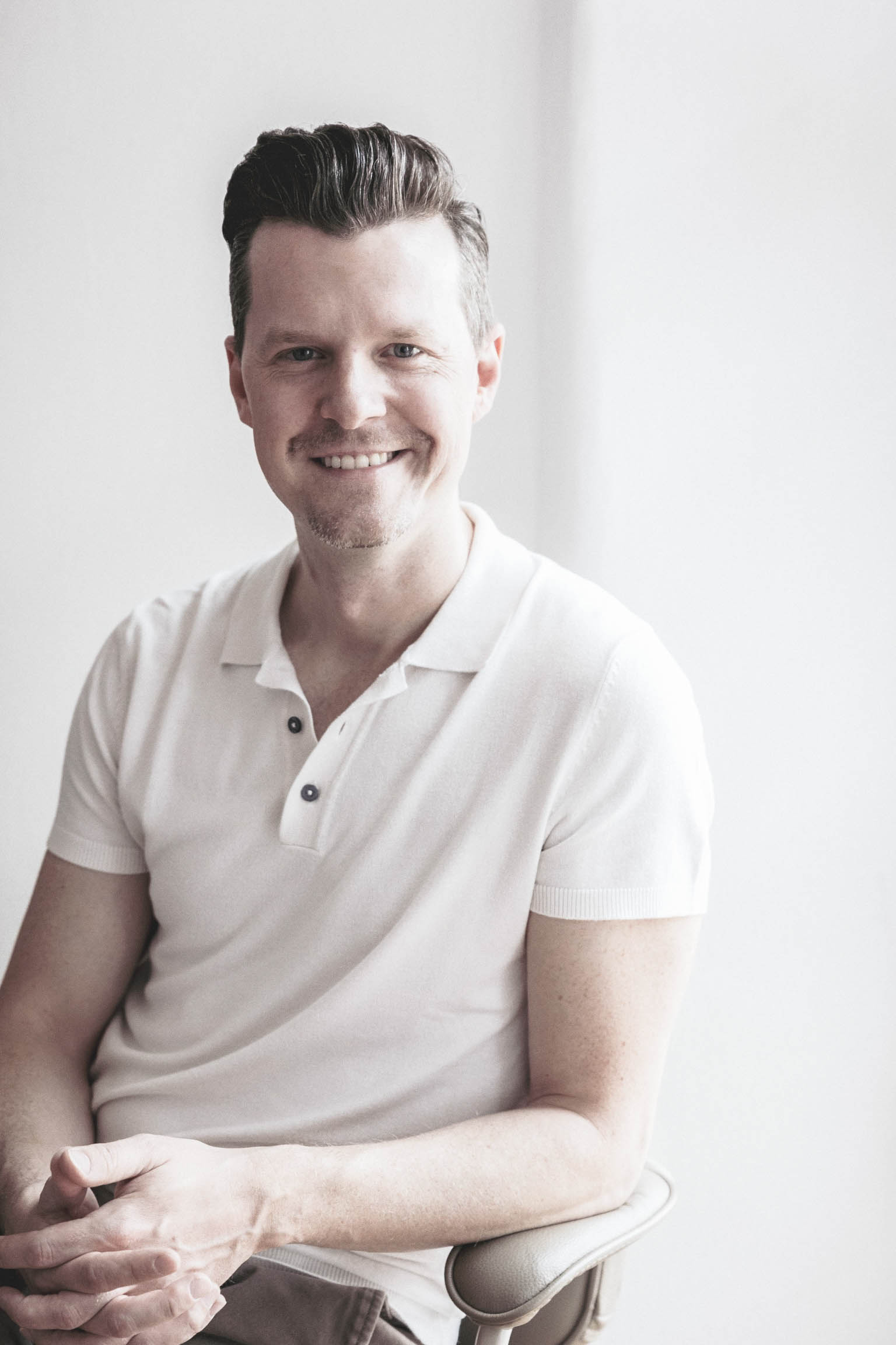 Ein Mann mit kurzen, dunklen Haaren sitzt auf einem Stuhl und lächelt in die Kamera. Er trägt ein weißes, kurzärmeliges Poloshirt und hat die Hände im Schoß gefaltet. Der helle, sanft beleuchtete Hintergrund vermittelt die einladende Atmosphäre des Creative Transformation Summit.