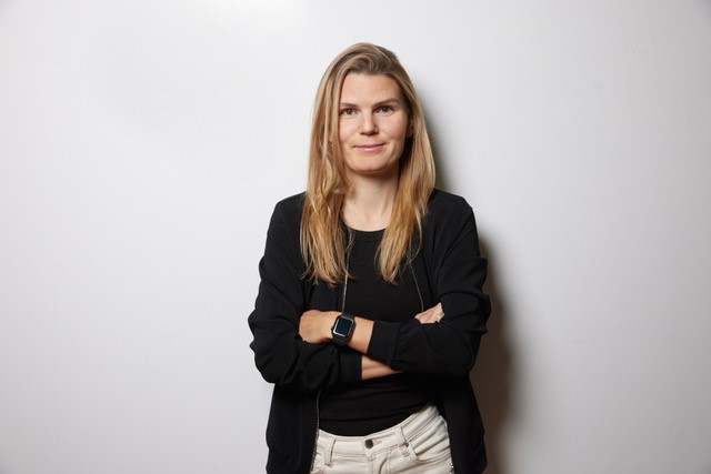 Eine Frau mit langen blonden Haaren steht mit verschränkten Armen vor einer schlichten weißen Wand. Sie trägt eine schwarze Jacke, ein schwarzes Hemd, eine weiße Hose und eine Smartwatch – ihre selbstbewusste Haltung erinnert an eine Hauptrednerin beim Creative Transformation Summit –, während sie mit einem leichten Lächeln in die Kamera blickt.