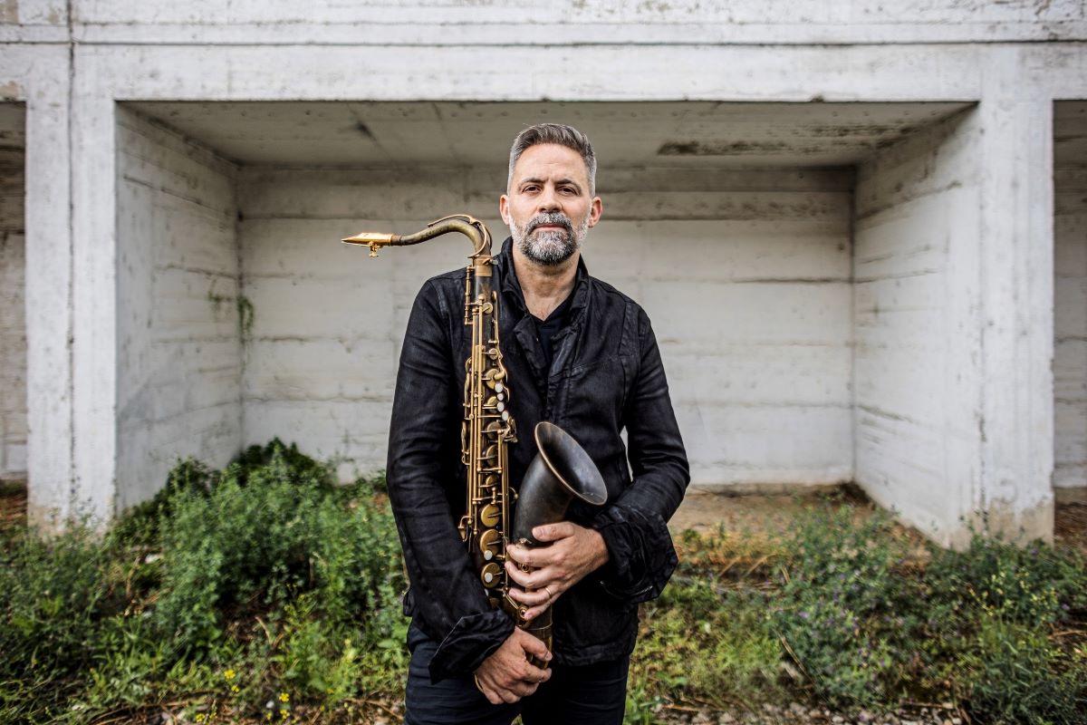Ulrich Drechsler, ein Musiker mit kurzem Haar und Bart, steht draußen vor einer Betonstruktur. In einer dunklen Jacke hält er sein Saxophon und ist von etwas Grün umgeben - bereit, neue Systeme zu komponieren.