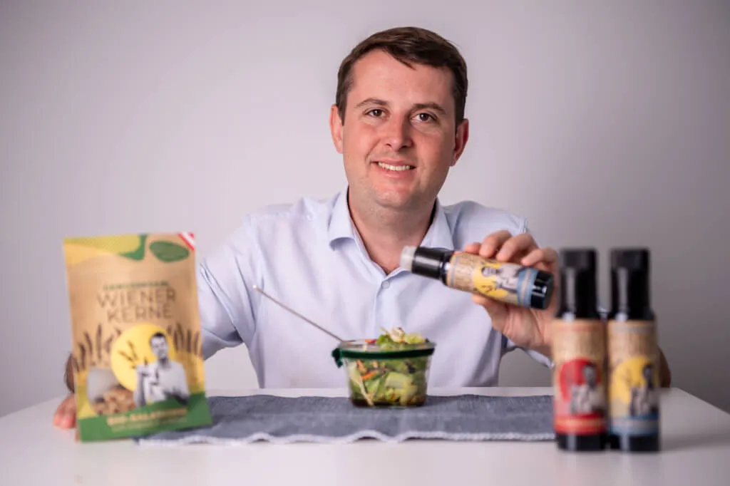 Ein Mann in einem weißen Hemd sitzt an einem Tisch, lächelt und hält eine Flasche Gewürz über einen Salat. Auf dem Tisch stehen zwei weitere Flaschen und ein Paket mit der Aufschrift "Wiener Kerne".