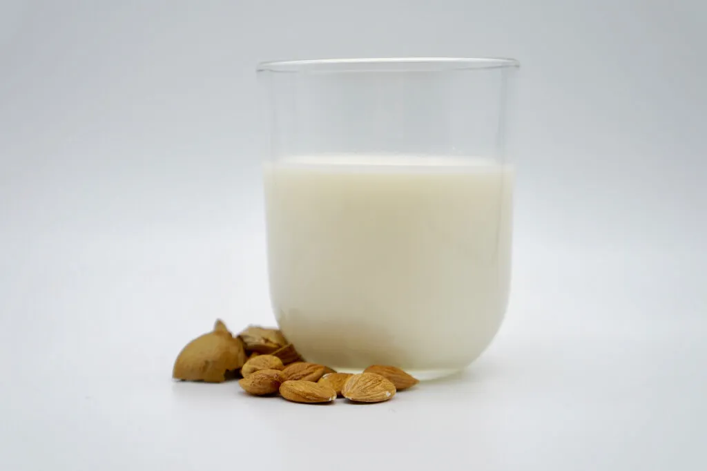 Ein klares, mit Mandelmilch gefülltes Glas steht auf einer weißen Fläche, daneben liegen ganze Mandeln und Mandelschalen.