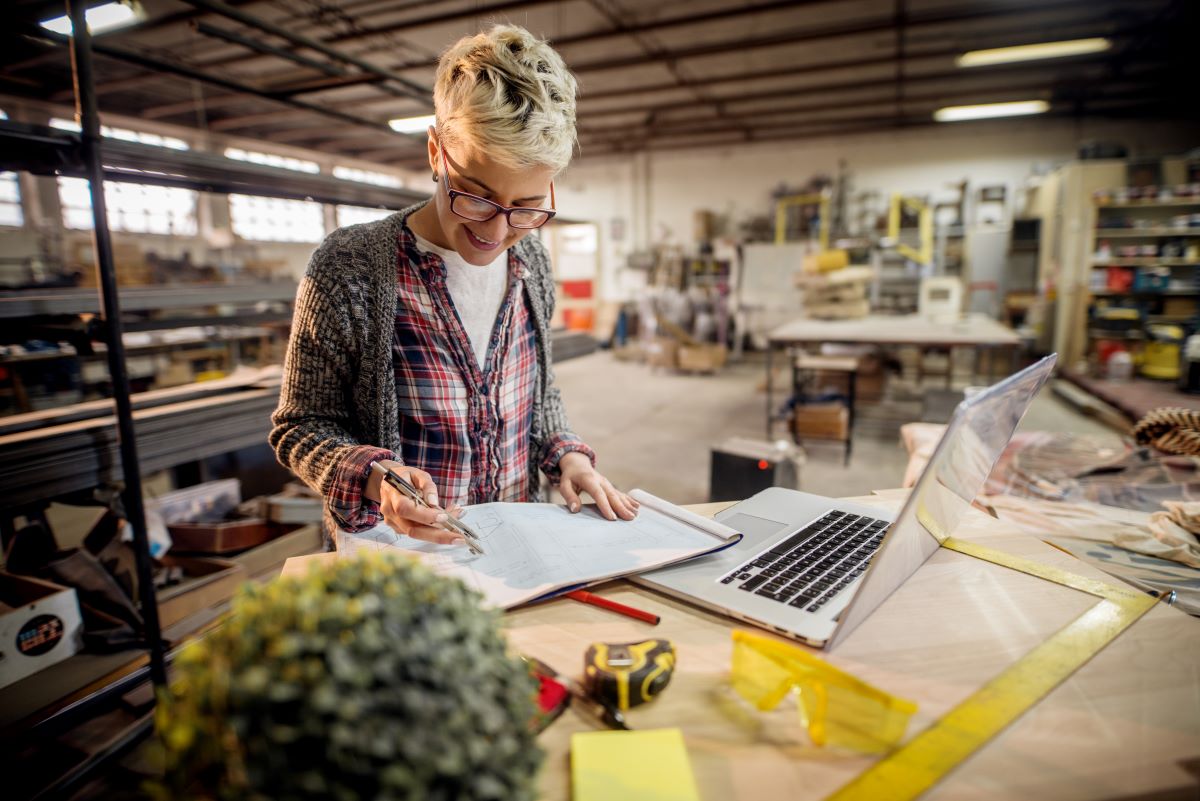 A person with short hair and glasses is working in a workshop. She is writing in a notebook while standing at a table with an open laptop, a tape measure, safety goggles and tools. Shelves and materials fill the background.