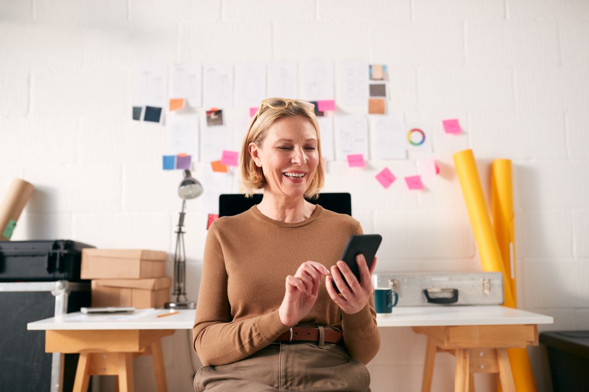 Eine Frau sitzt an einem Schreibtisch in einem B&uuml;ro, l&auml;chelt und benutzt ihr Smartphone - vielleicht nimmt sie an einer Umfrage &uuml;ber die Wiener Kreativwirtschaft teil. Hinter ihr stehen B&uuml;romaterialien, Kisten und Papiere mit Klebezetteln, die an eine wei&szlig;e Wand gepinnt sind.