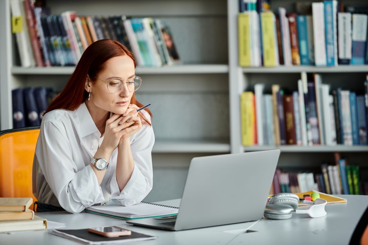 Eine Frau mit Brille sitzt an einem Schreibtisch vor einem Laptop, hält einen Bleistift in der Hand und blickt auf den Bildschirm, während sie an einer Umfrage zur Wiener Kreativwirtschaft teilnimmt. Auf dem Schreibtisch liegen Bücher, Kopfhörer und Büromaterial, im Hintergrund sind Bücherregale zu sehen.