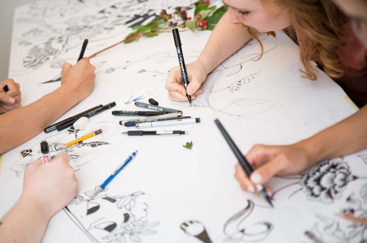 Drei Personen von Illustration Ladies in Wien zeichnen mit Stift und Haltung detaillierte, florale und abstrakte Motive mit schwarzen Markern und Stiften auf ein großes Blatt Papier, auf dem verschiedene Kunstmaterialien verstreut liegen.