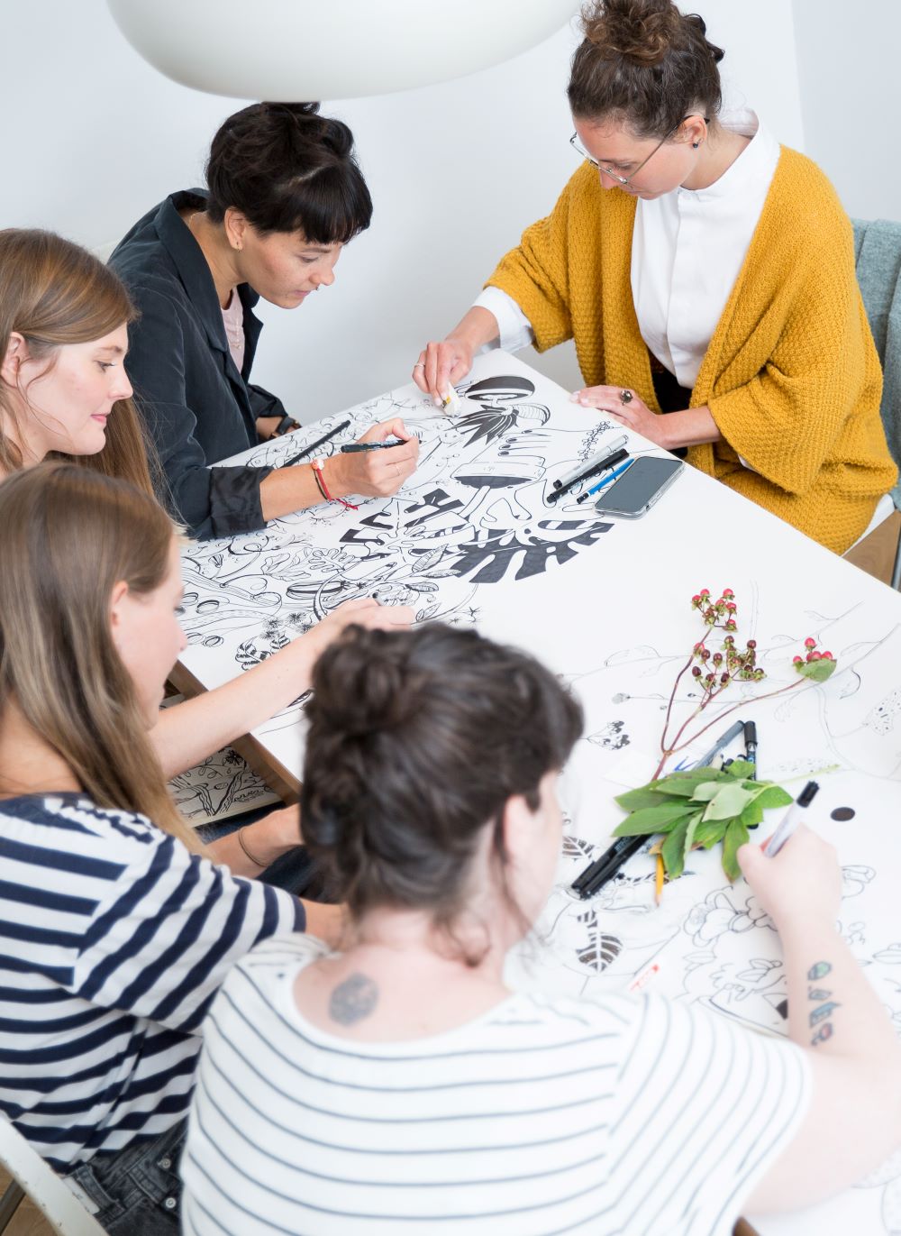 Fünf Illustration Ladies sitzen in Wien um einen Tisch und arbeiten gemeinsam an einer großen Schwarz-Weiß-Zeichnung. Stift und Haltung leiten ihren kreativen Prozess, während Malutensilien und Blätter verstreut werden, was ihre Konzentration und ihr Engagement widerspiegelt.
