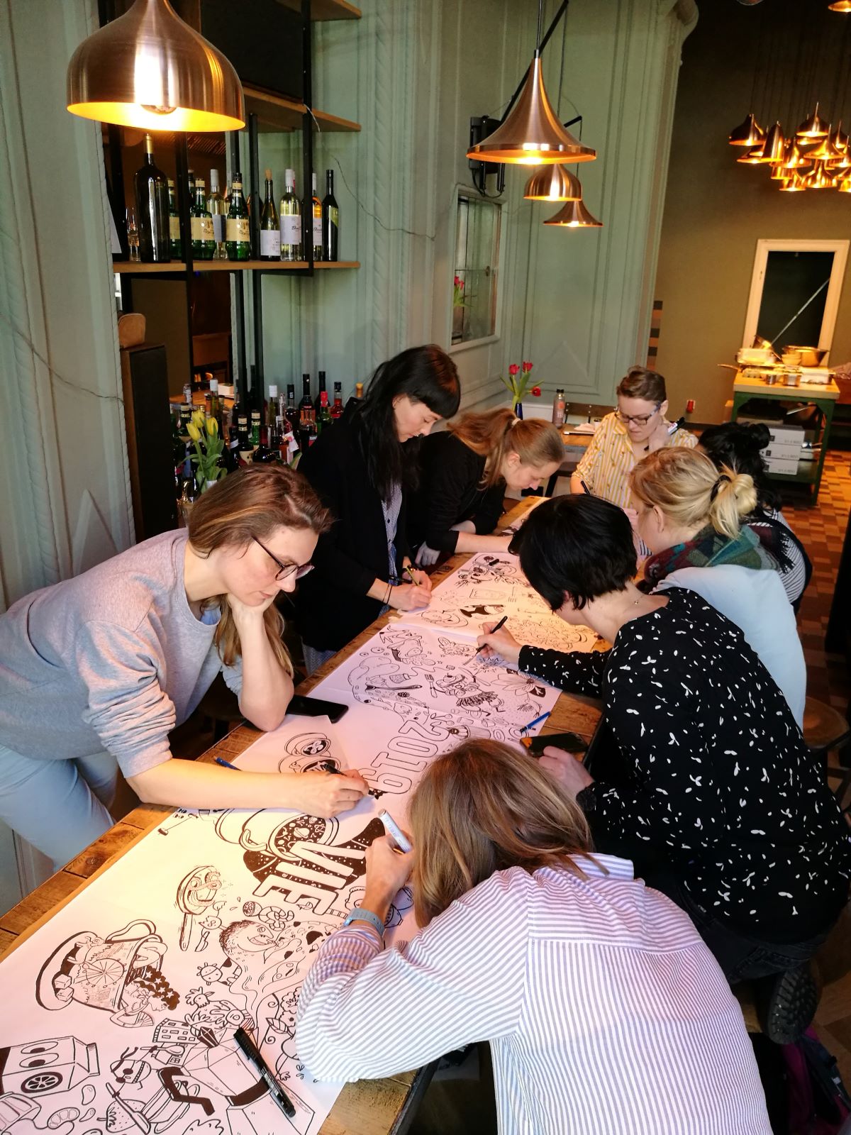 In einem gemütlichen, warm beleuchteten Raum mit Hängelampen und Flaschen in Regalen im Hintergrund ist eine Gruppe von Menschen um einen Tisch in Wien versammelt und malt große Illustration Ladies-Plakate mit Stiftmarkern aus.